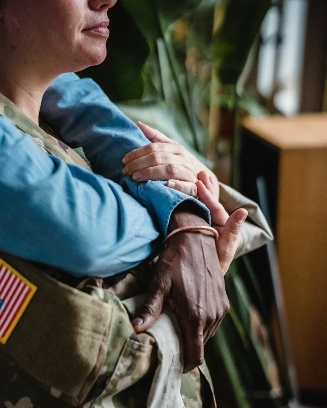 Close up of a woman in uniform, her partner is has arms around her from behind though we can't see the partner's face. They are considering discernment counseling for their relationship and want to proceed with kindness for all involved.