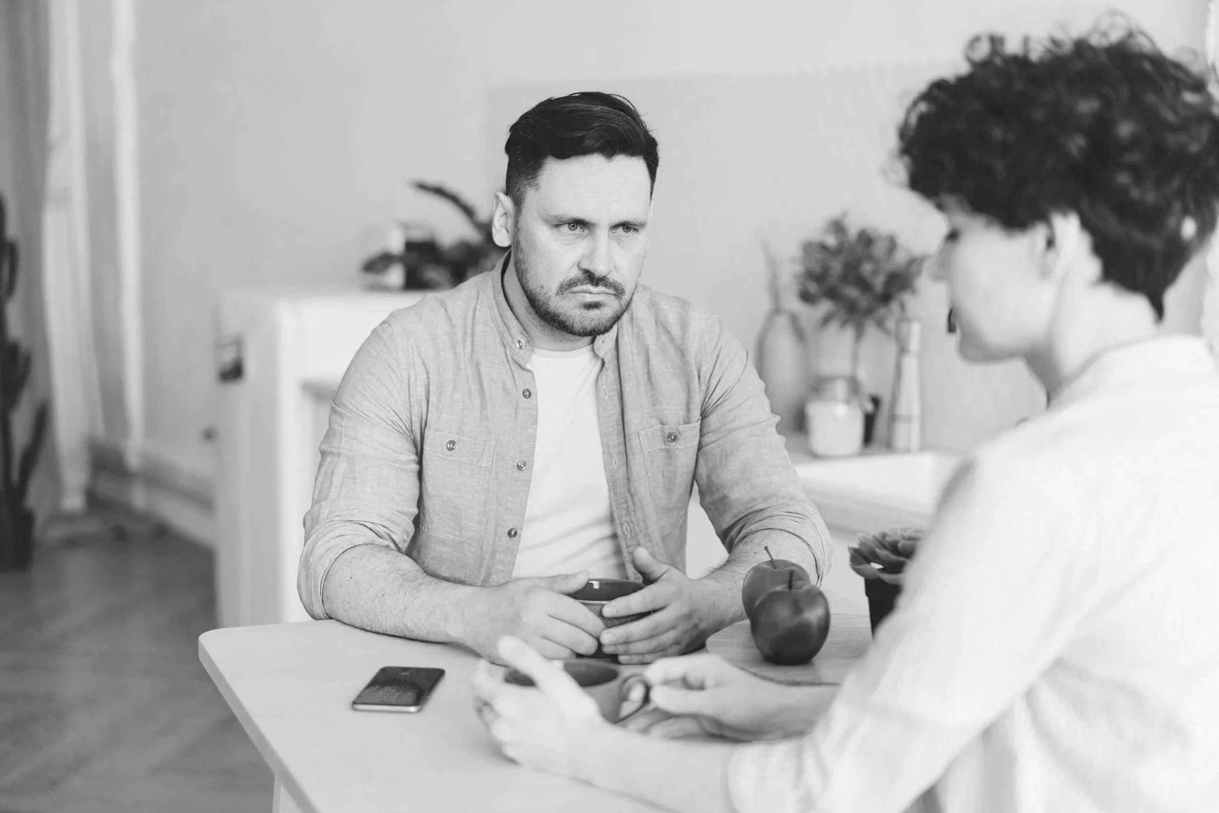 Man sitting at a table with a family member looking angry as he's hearting the other person talk. They have dishes and food between them, at home. They are discussing family counseling in Riverside with Inland Empire Couples Counseling.