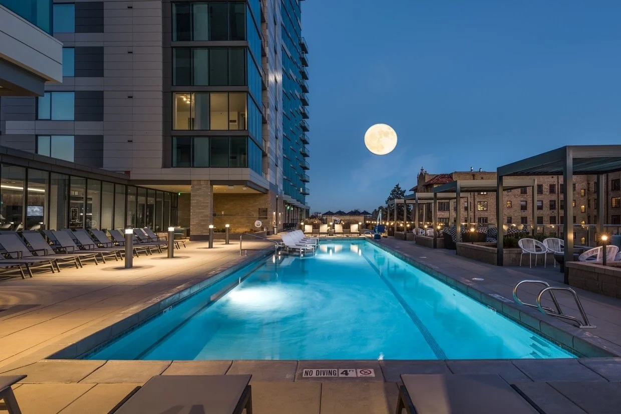 City apartment building rooftop pool area at night with lounge chairs, outdoor seating, and a view of a full moon in the sky.