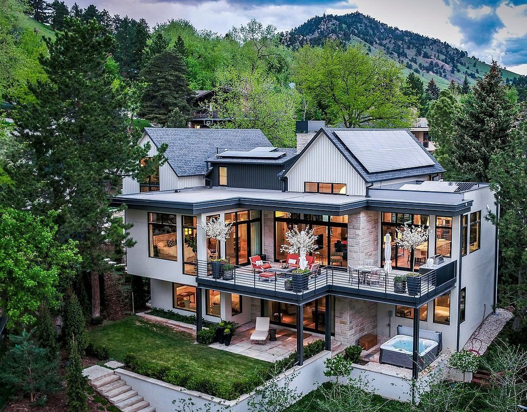 Modern two-story house with large glass windows and a balcony, surrounded by trees and mountains in the background.