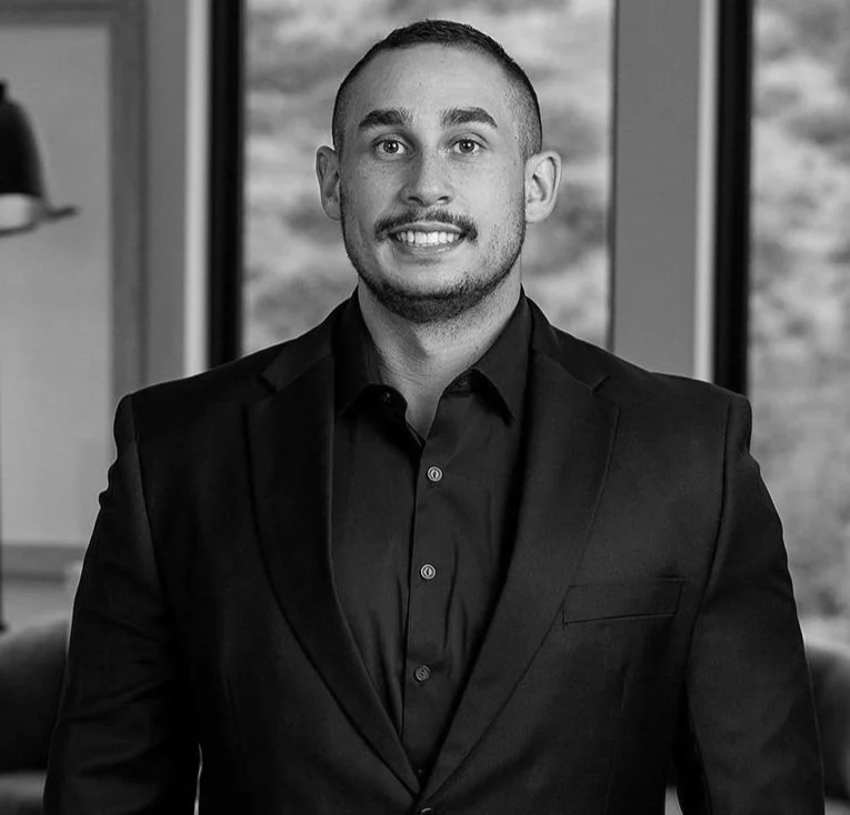A man in a black suit and shirt smiling in an indoor setting with large windows and outdoor scenery in the background.