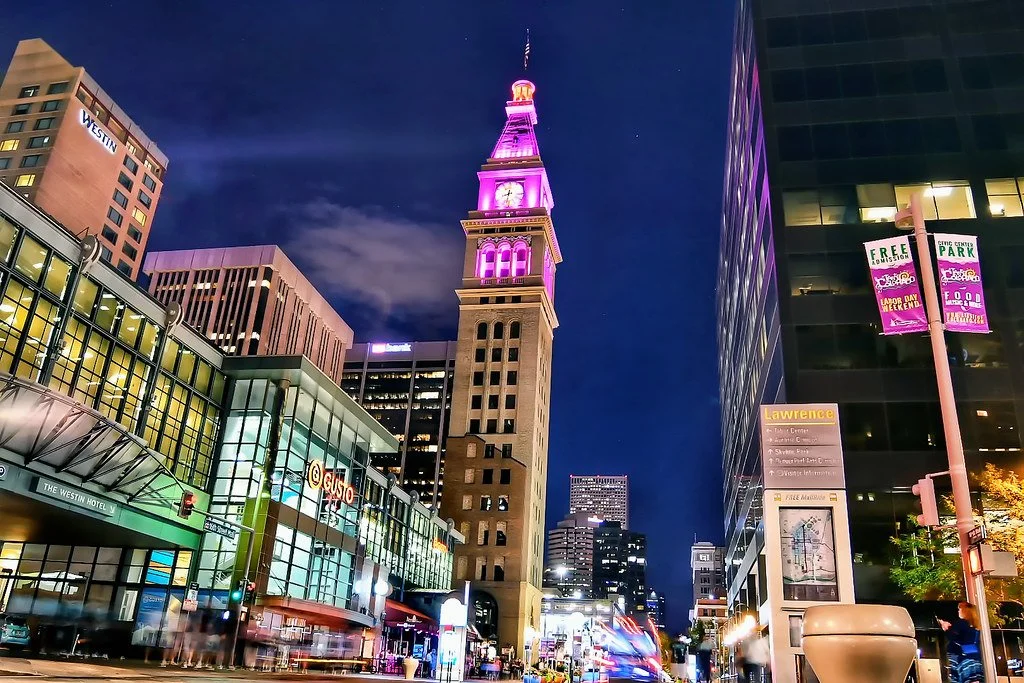 Nighttime city street scene with tall clock tower illuminated in pink and purple lights, surrounded by modern high-rise buildings and busy traffic. The Westin hotel sign is visible on a building to the left.