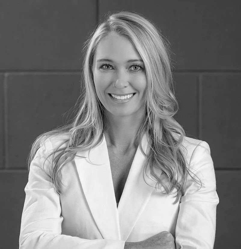 A smiling woman with long wavy hair wearing a white blazer, posed with her arms crossed in front of a plain wall.
