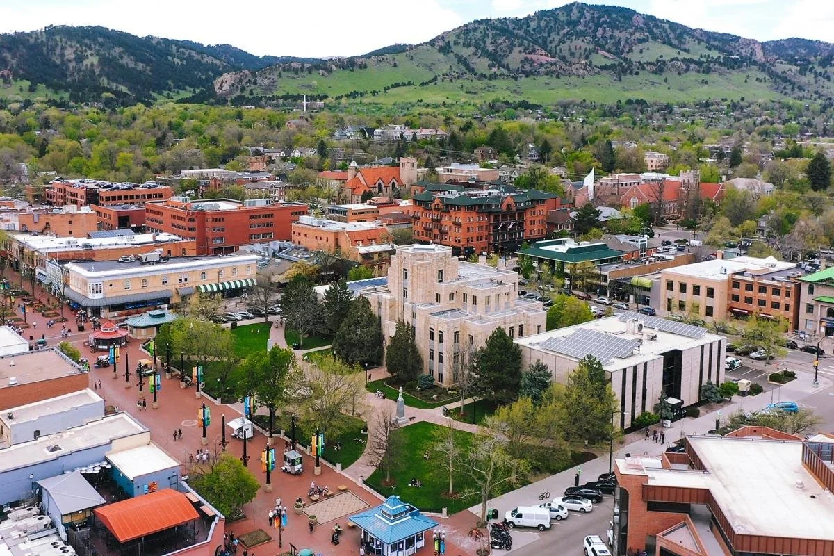 A view of a downtown area with brick buildings, trees, and mountains in the background, showing a lively street with pedestrians and parked cars.