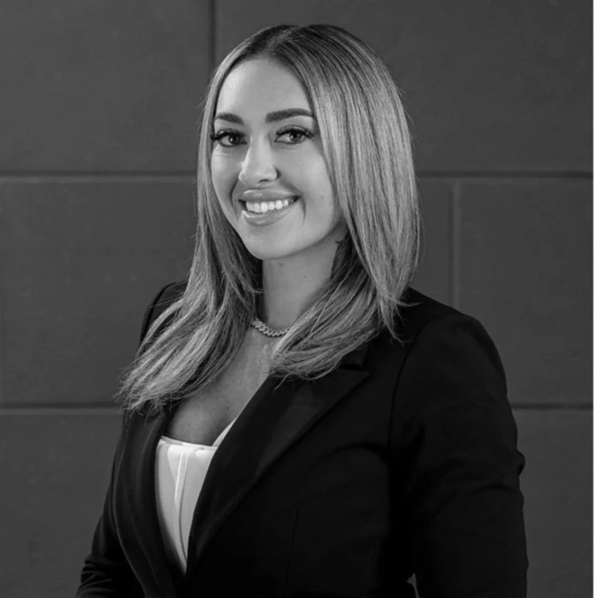 A woman with long, straight hair wearing a blazer and necklace, smiling at the camera against a plain background.