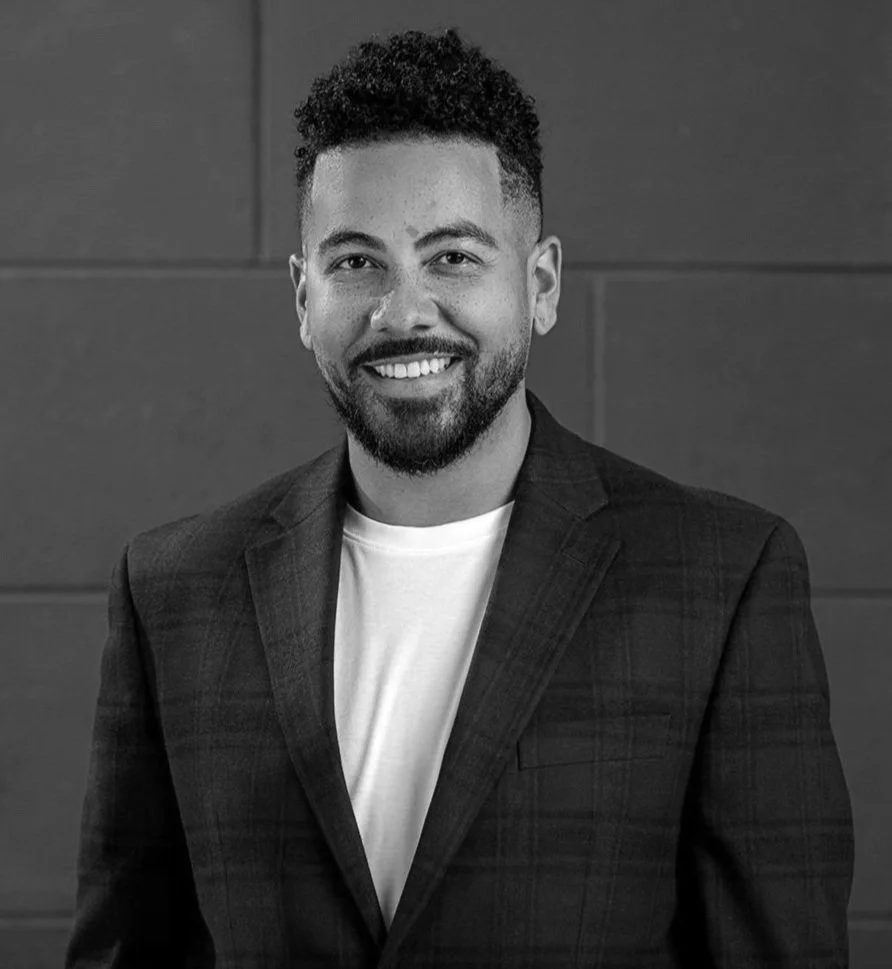 Black and white portrait of a man with short curly hair and a beard, wearing a blazer and T-shirt, smiling at the camera.