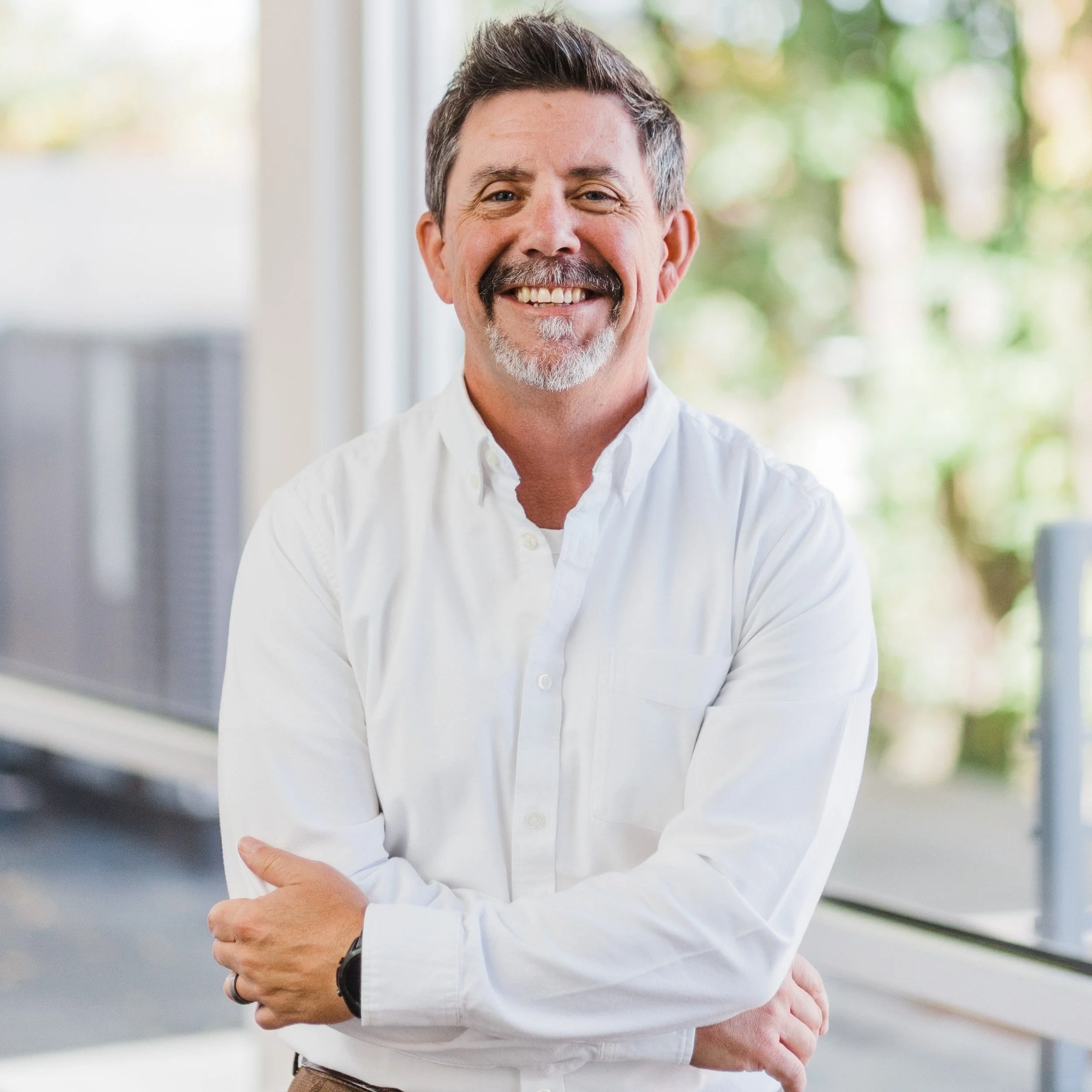 Smiling middle-aged man with a beard and mustache wearing a white button-up shirt standing with arms crossed in front of a window with green trees outside.