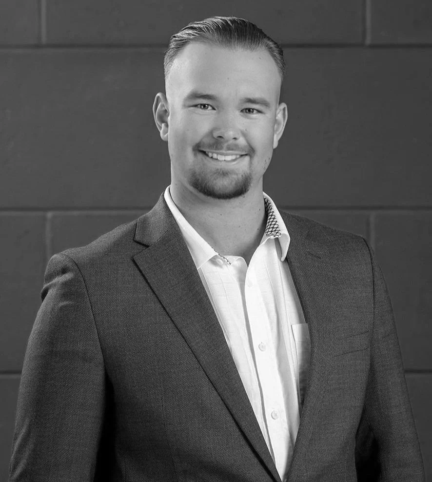 Black and white professional headshot of a smiling young man with short hair, dressed in a suit jacket and shirt, posing against a wall background.