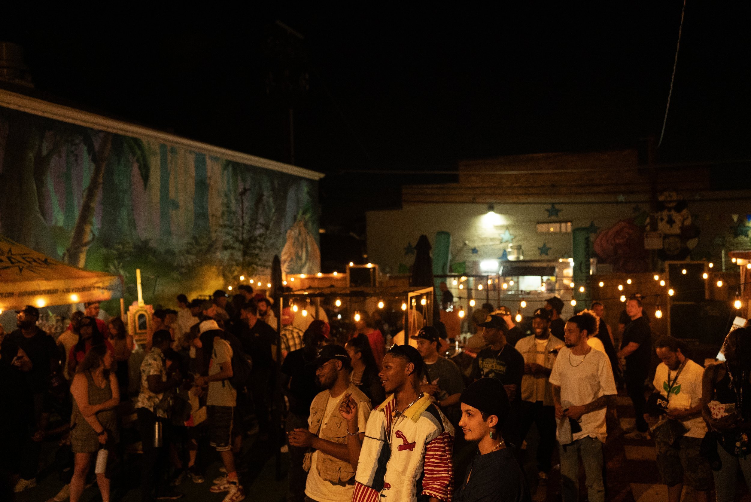 Nighttime outdoor event with a crowd of diverse people, string lights, and colorful murals on the walls.