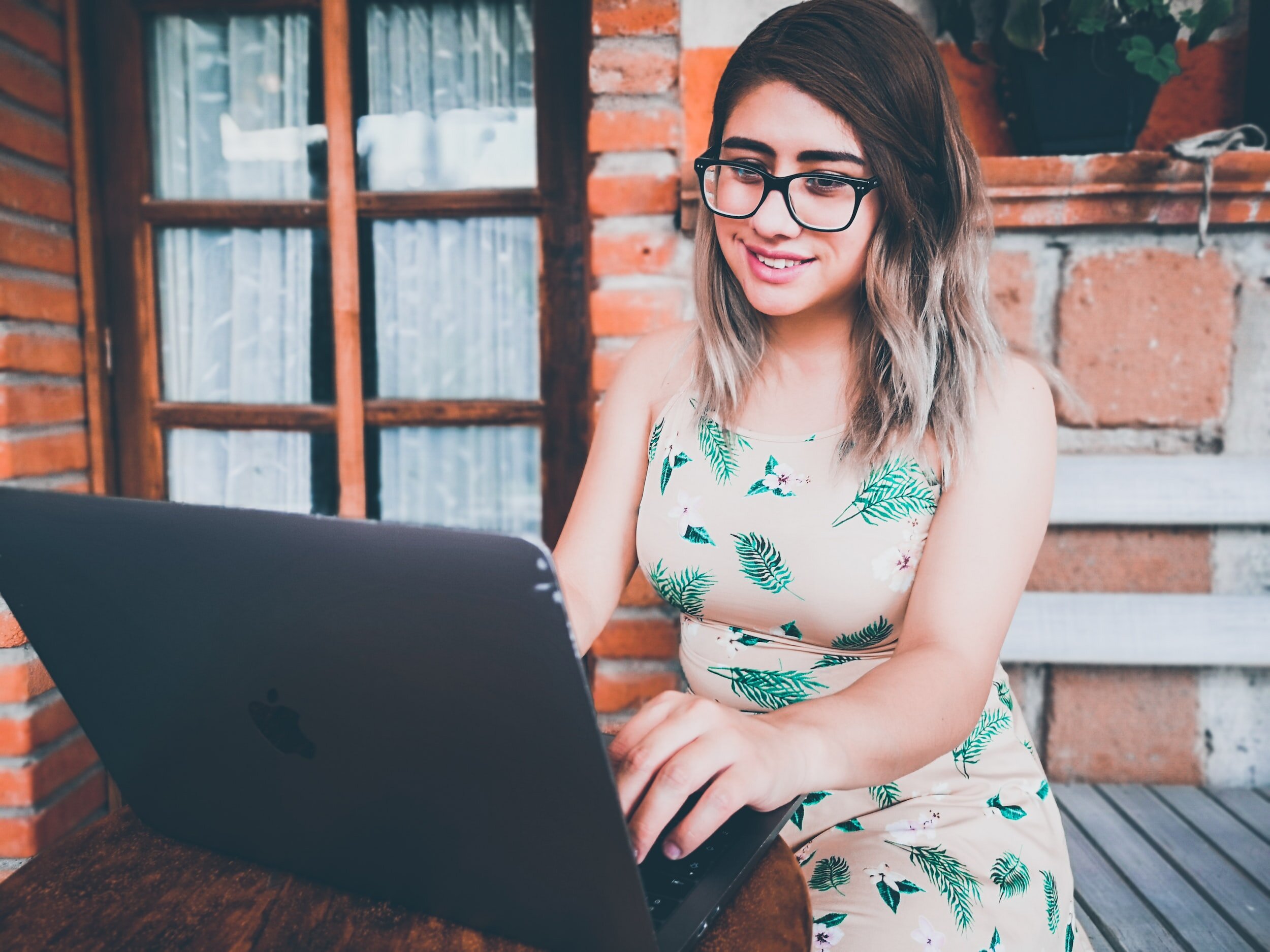 A young woman with wavy, shoulder-length hair and glasses smiling while working on a black laptop outdoors on a wooden patio, with a brick and window background.