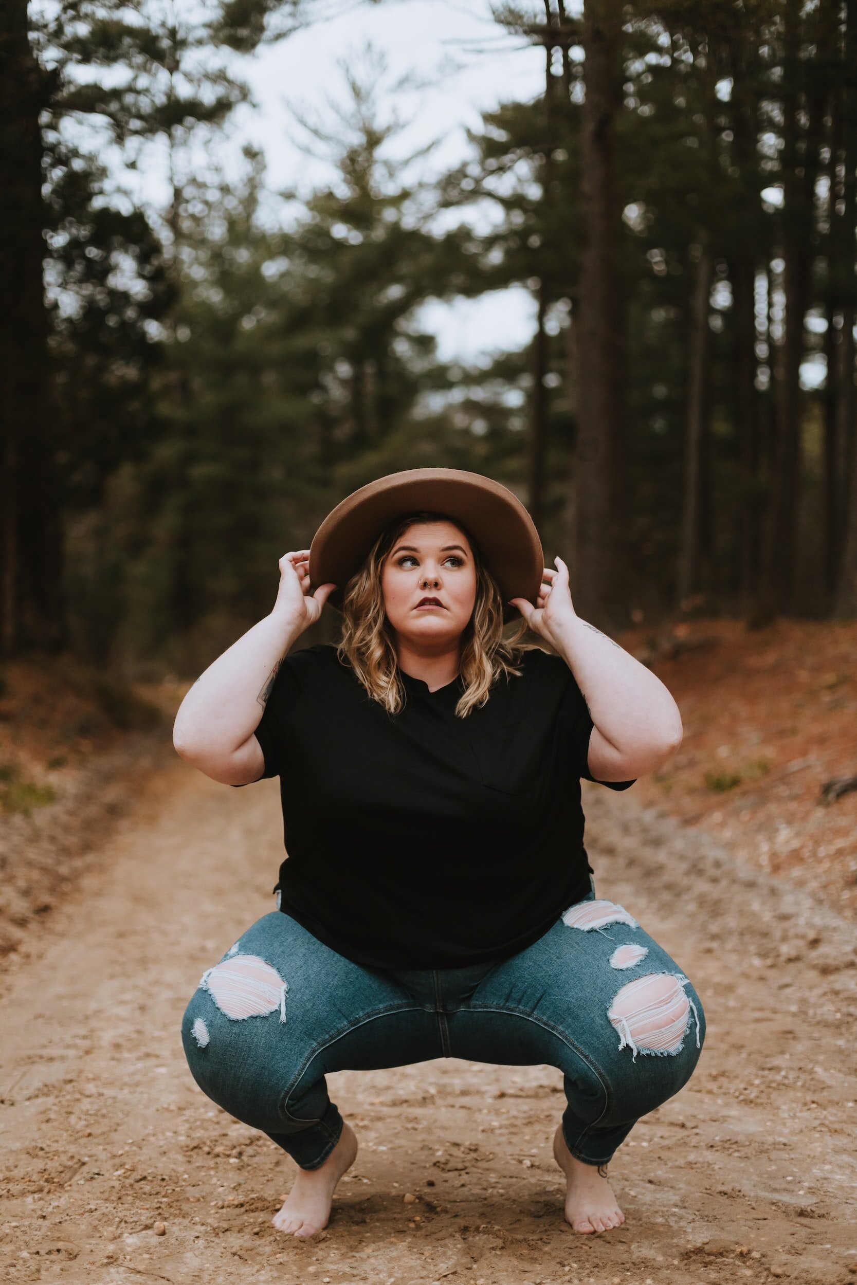 A woman squatting on a forest dirt path, holding a wide-brimmed brown hat, wearing a black shirt and ripped jeans.