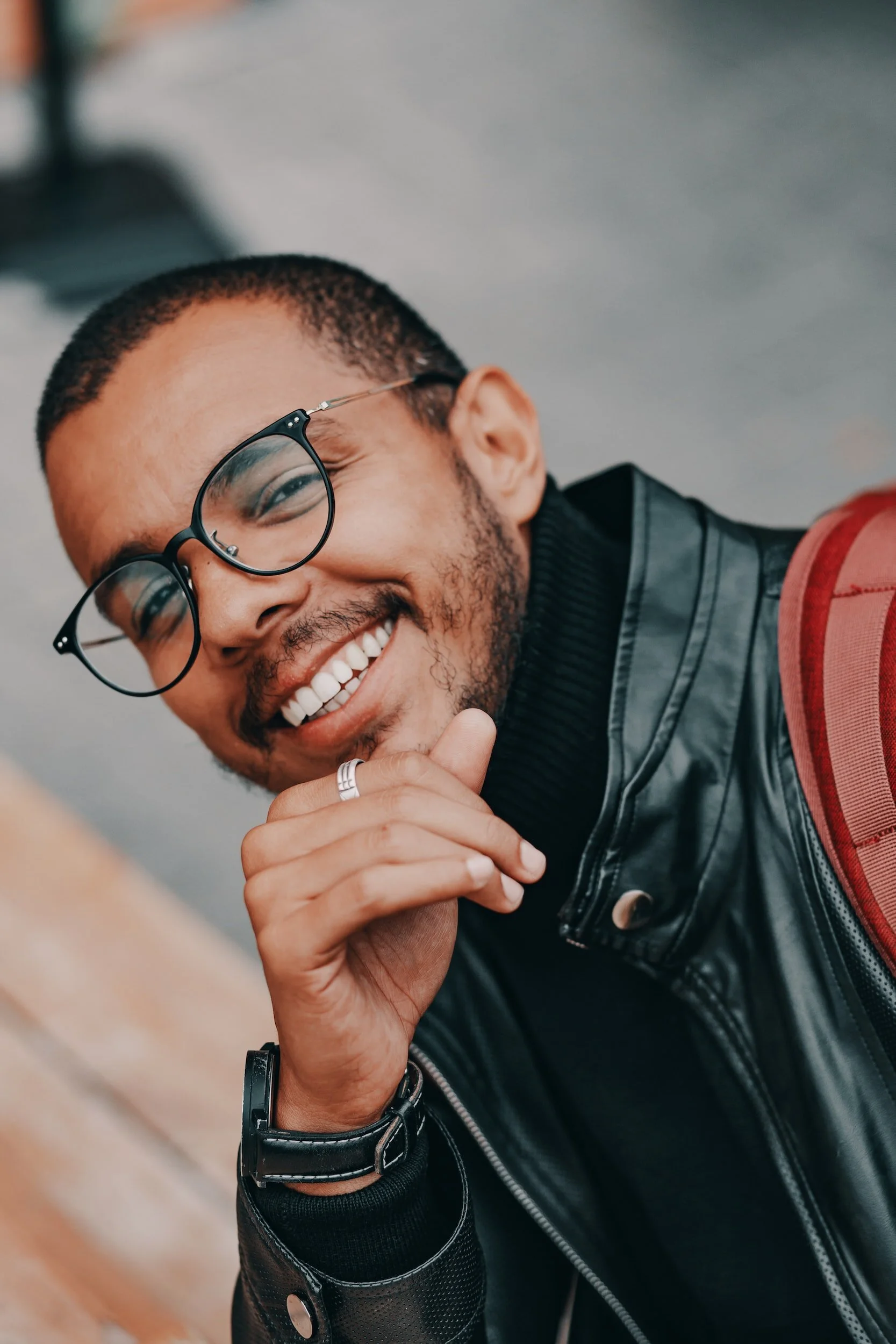 Smiling young man with glasses, wearing a black leather jacket, holding his chin, and carrying a red backpack.
