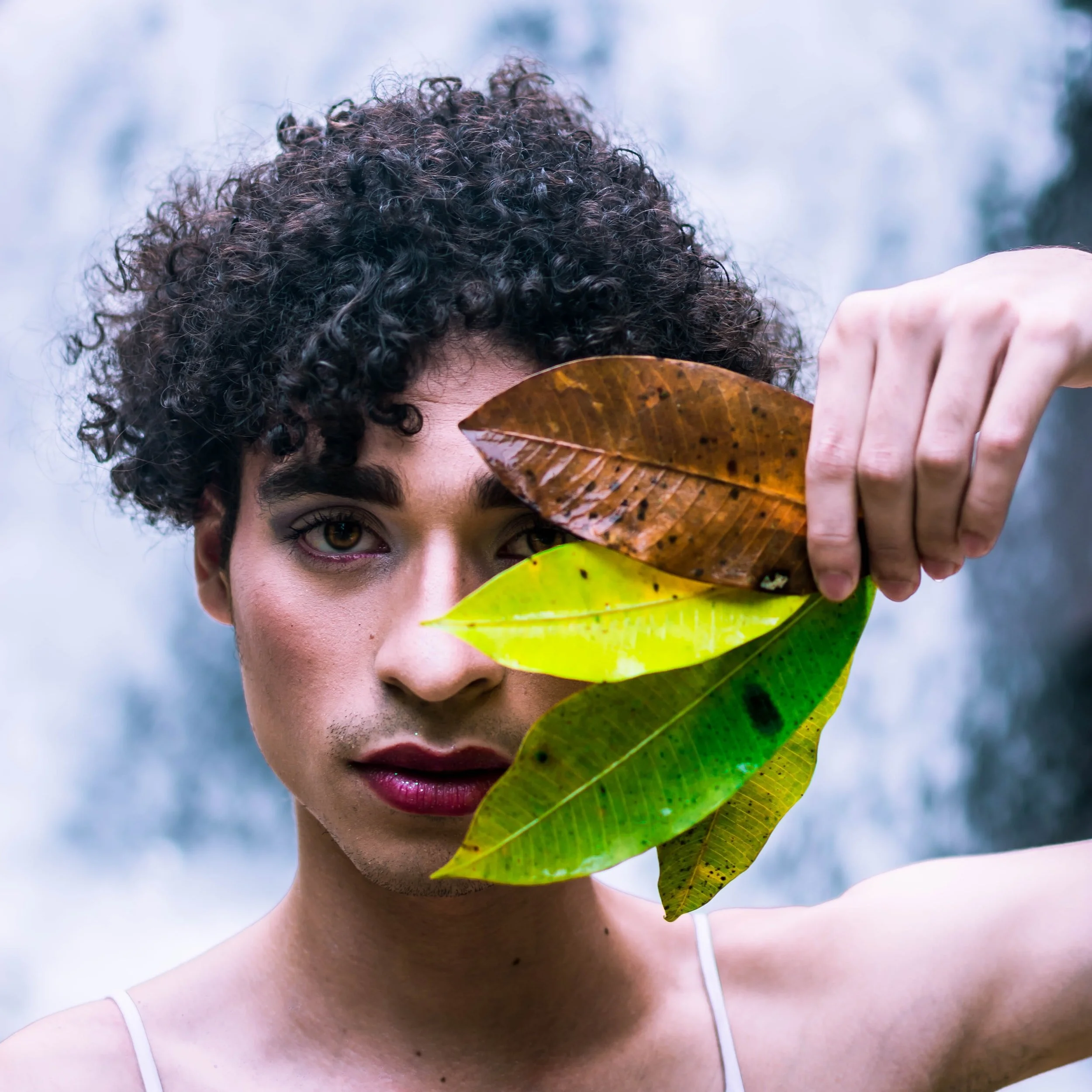A woman with curly hair holds a bunch of colorful leaves in front of her face, partially covering one eye.
