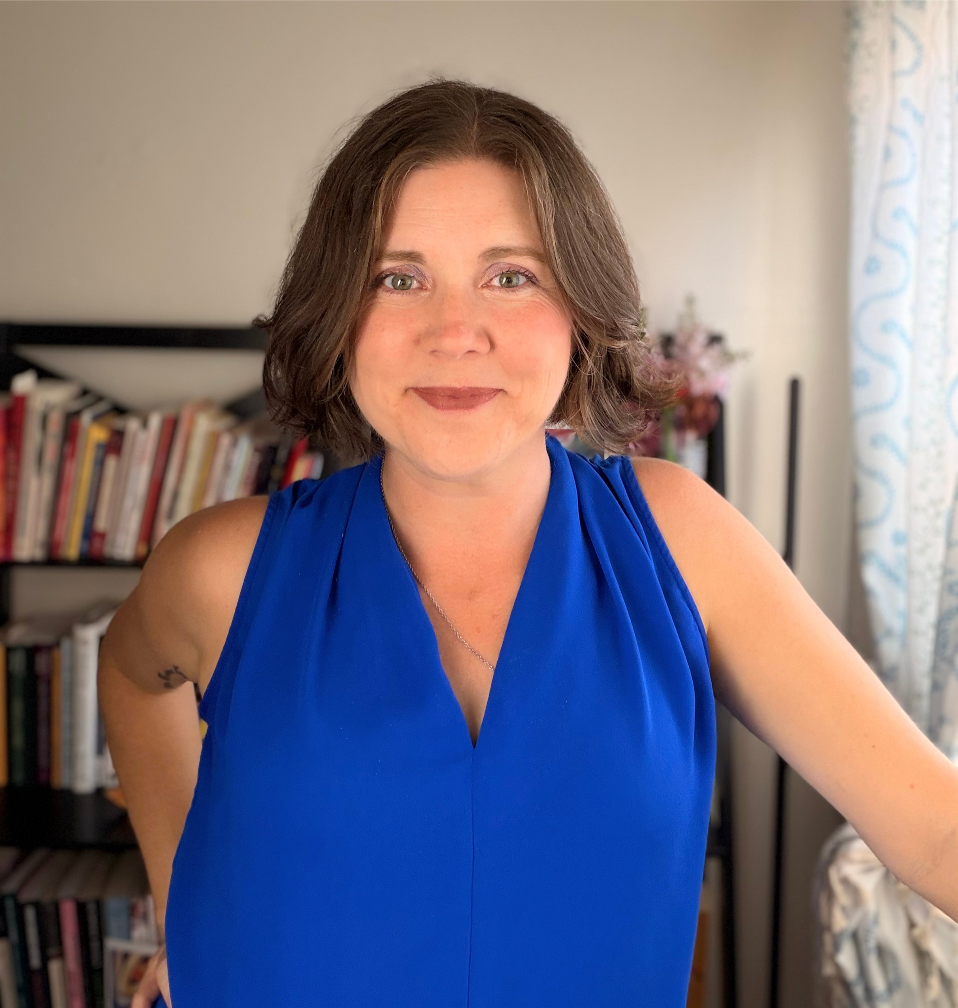 A woman with shoulder-length brown hair wearing a blue sleeveless top, smiling, with a bookshelf and a window with patterned curtains in the background.