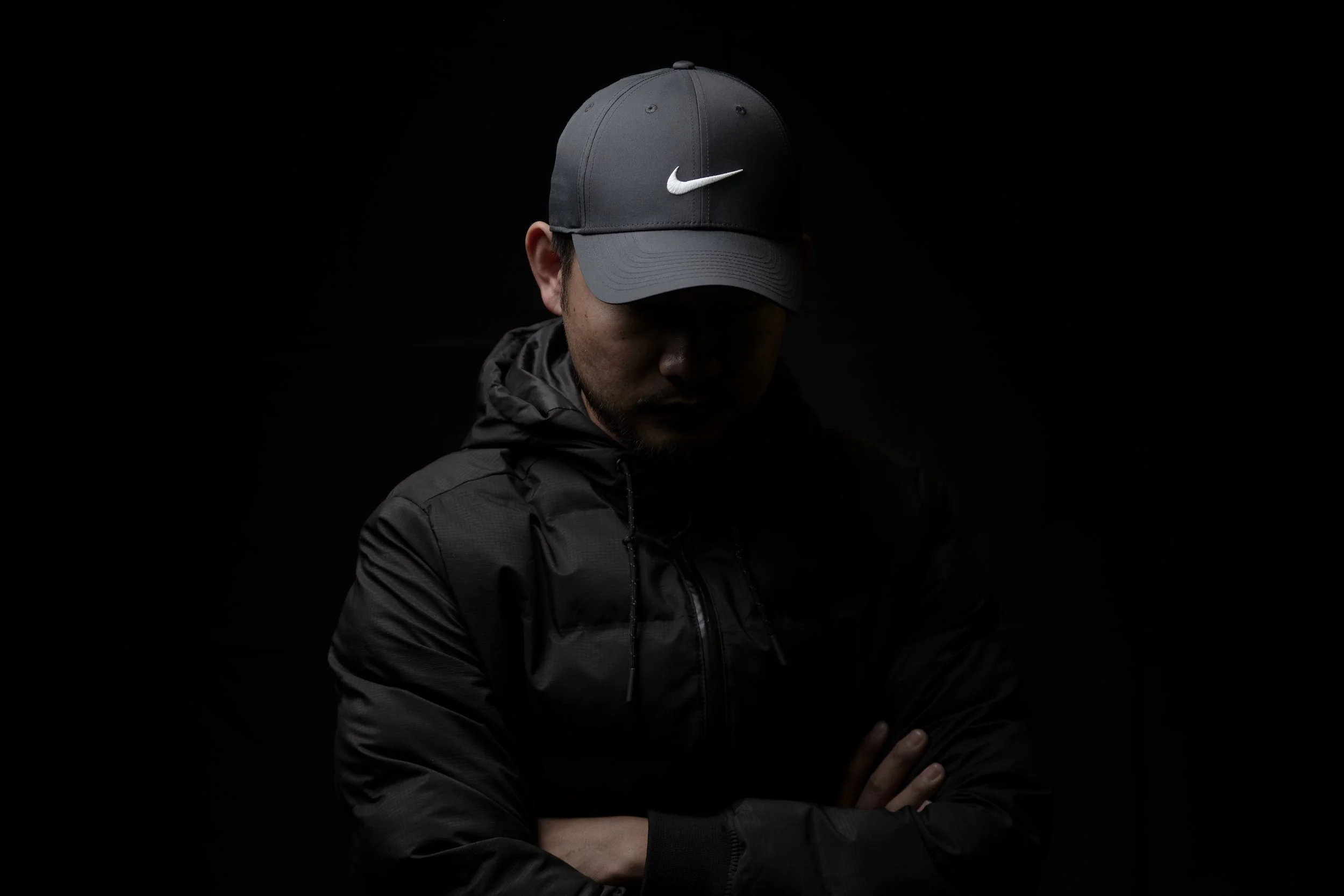 A man wearing a dark gray Nike cap and a black jacket, standing against a black background with his head slightly down and arms crossed.