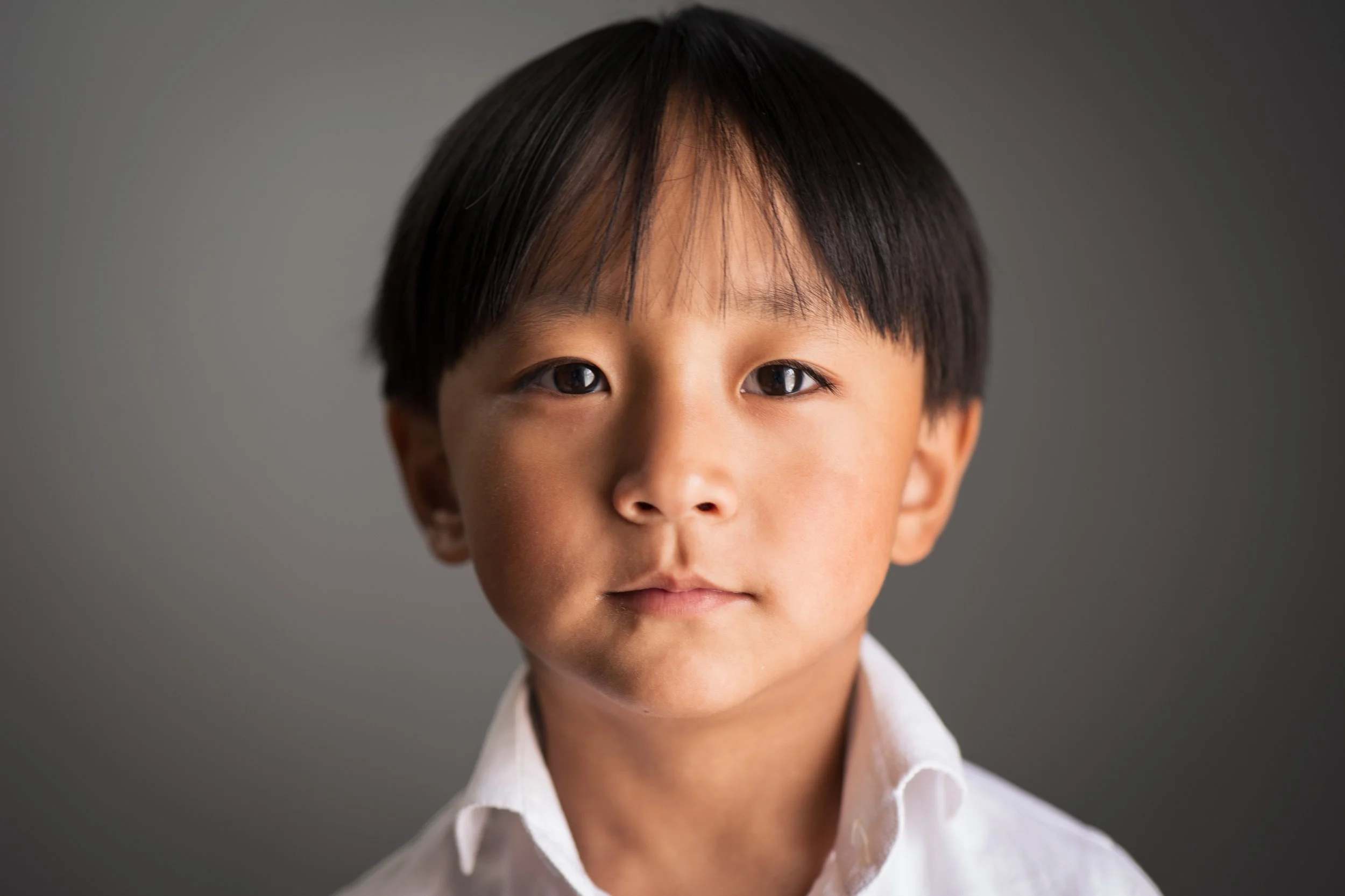A young boy with dark hair and brown eyes, wearing a white collared shirt, looking directly at the camera against a gray background.