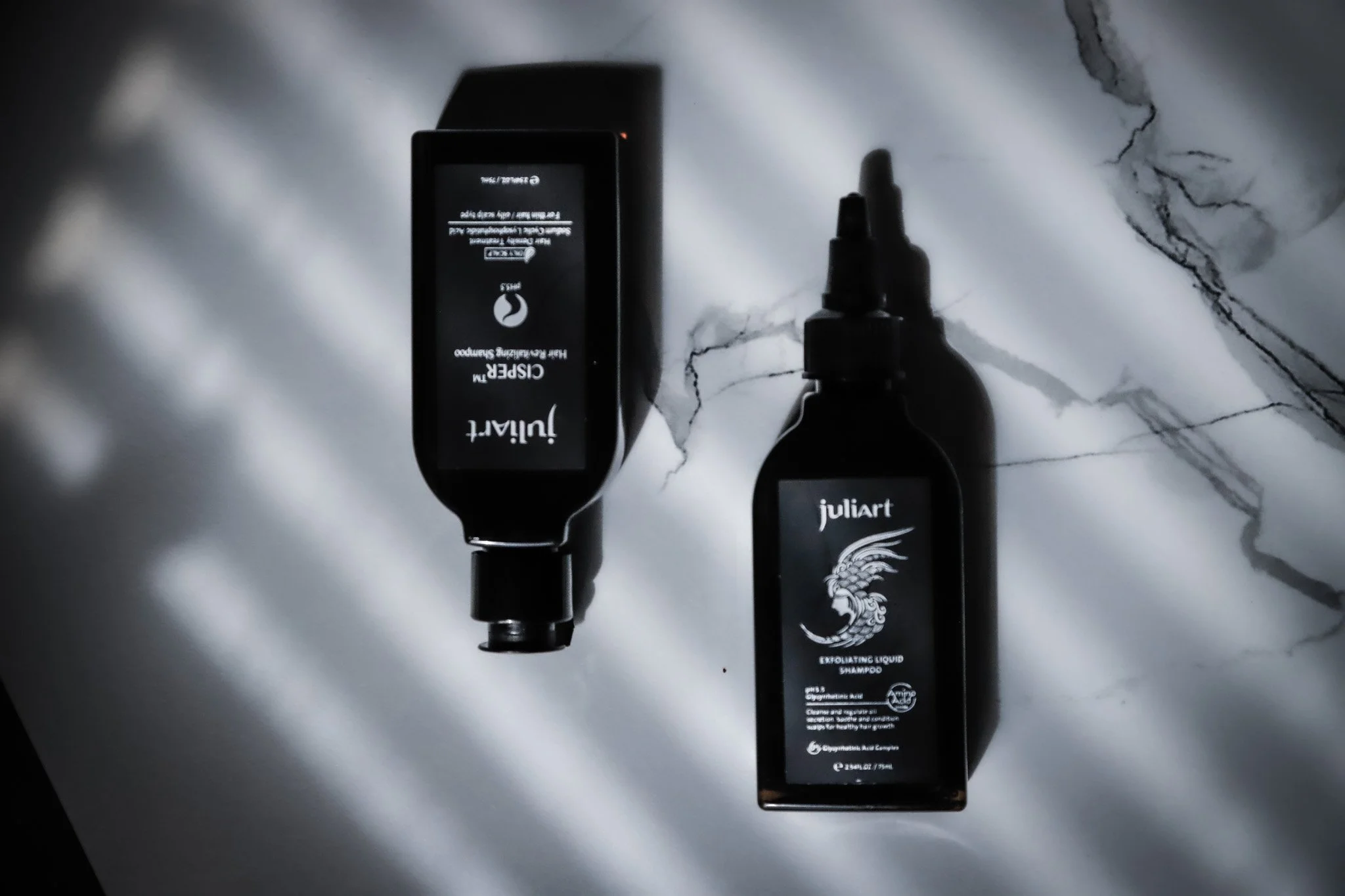 Two black bottles of Juliart exfoliating liquid shampoo with black labels, placed on a white marble surface with black veins, casting shadows.