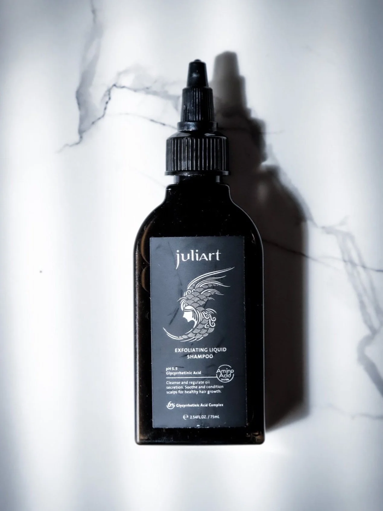 A black bottle of Juliart exfoliating liquid shampoo with a spray top, placed on a white marble surface.