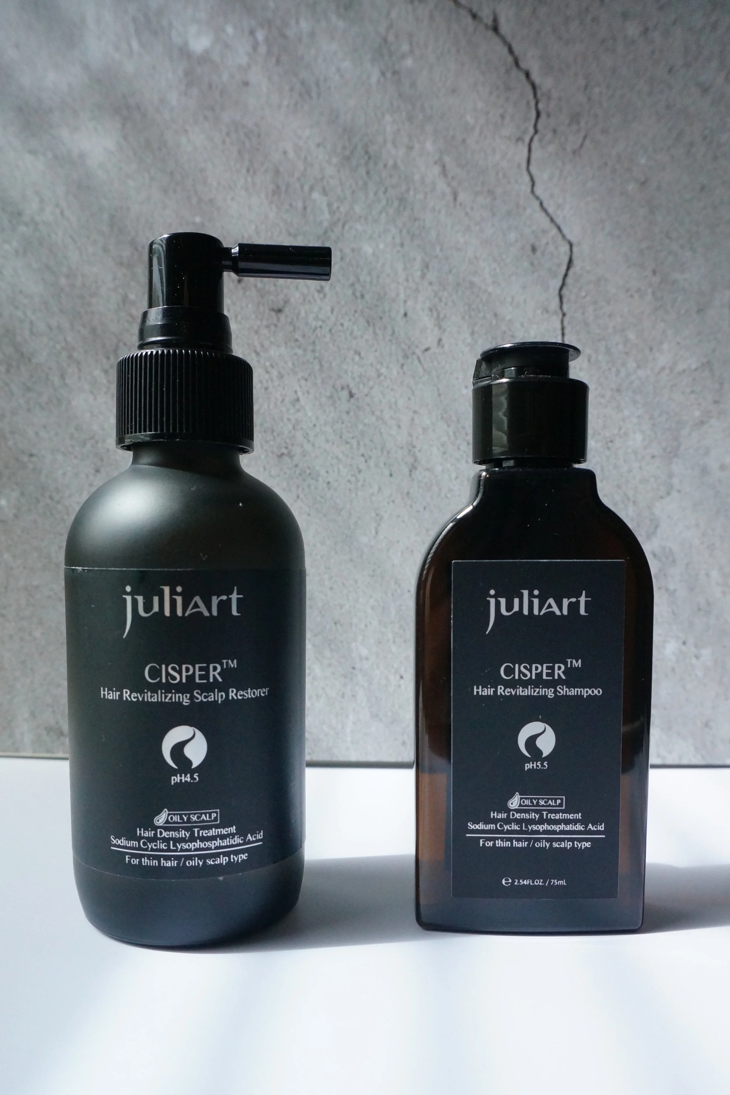Two bottles of Juliart Cisper Hair Revitalizing scalp and shampoo treatment for oily scalps, with black labels and pumps, placed on a white surface against a gray cracked wall background.