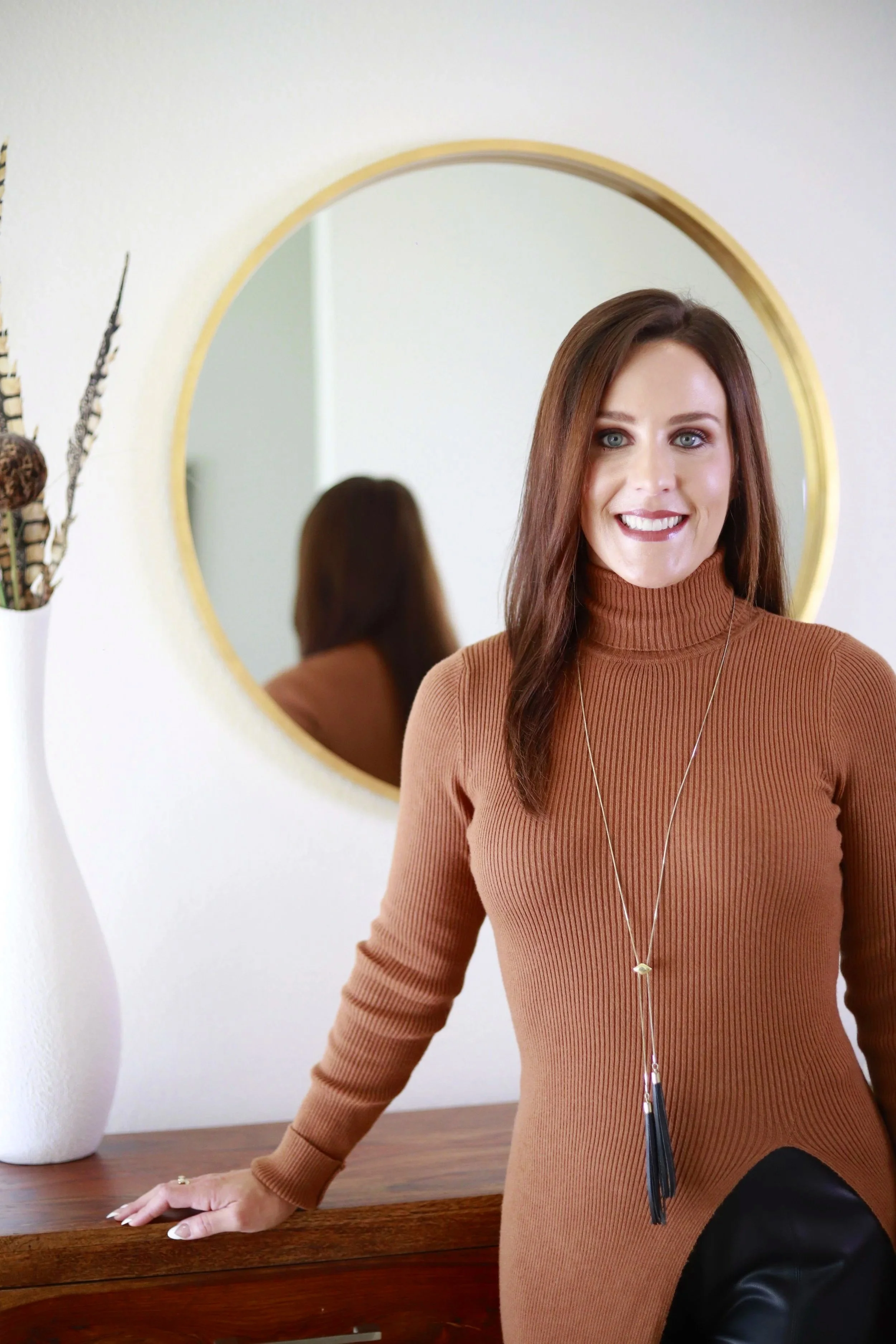 A woman with brown hair and blue eyes wearing a brown turtleneck sweater and long necklace, standing in front of a mirror with a gold frame, smiling at the camera, with a white vase and dried plants on the side.