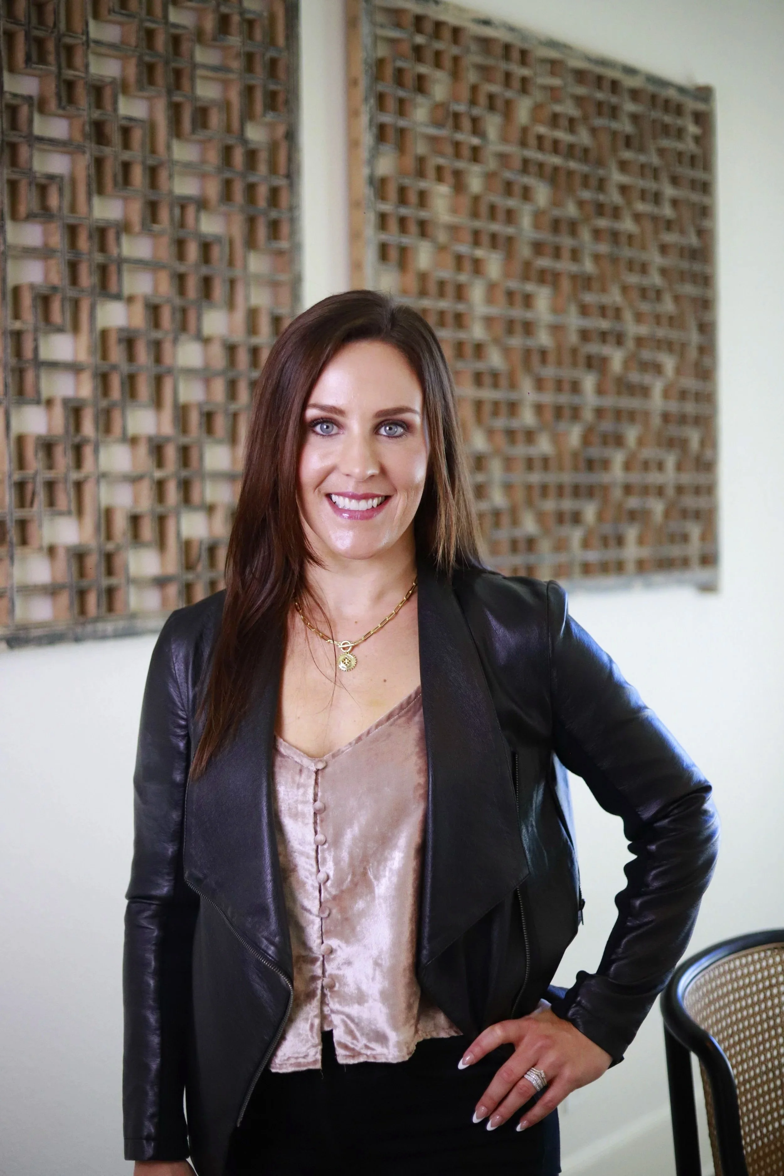 A woman with long brown hair, blue eyes, and a bright smile, wearing a black leather jacket, a light pink satin blouse, gold jewelry, and black pants, standing indoors in front of a decorative wall with wooden lattice panels.