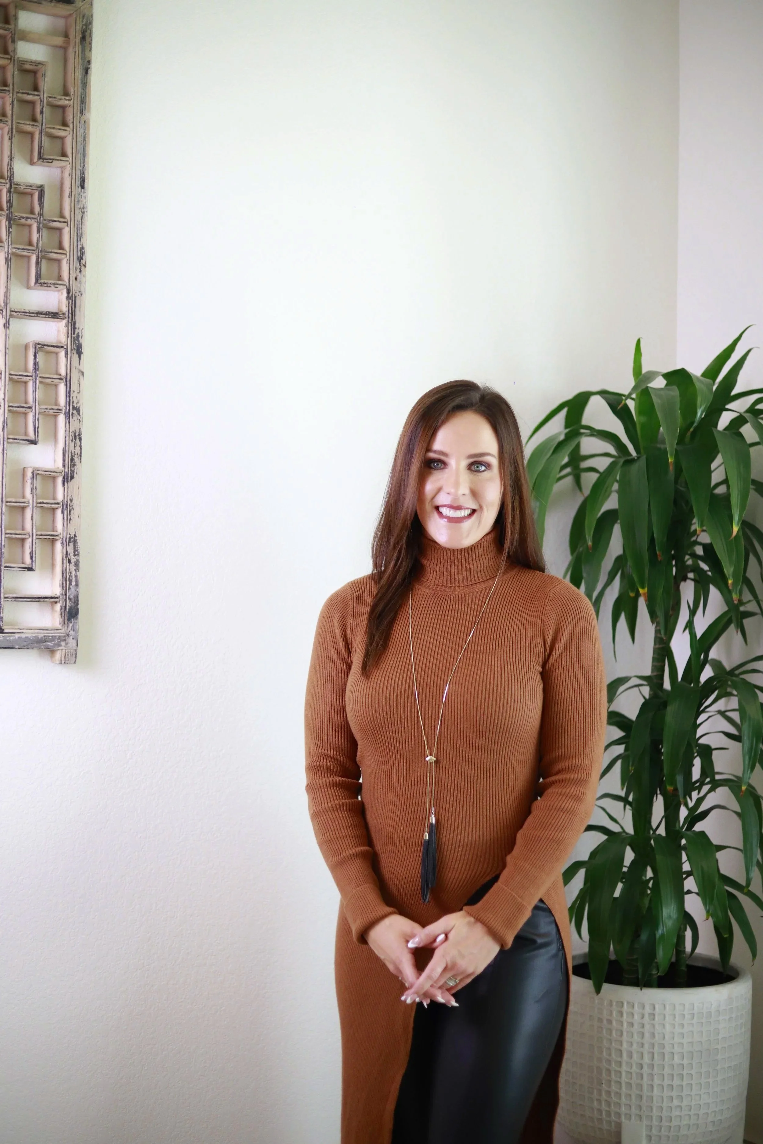 A woman with long brown hair, wearing a brown turtleneck sweater and a long necklace with a black tassel, stands in front of a white wall and a large green potted plant, smiling.