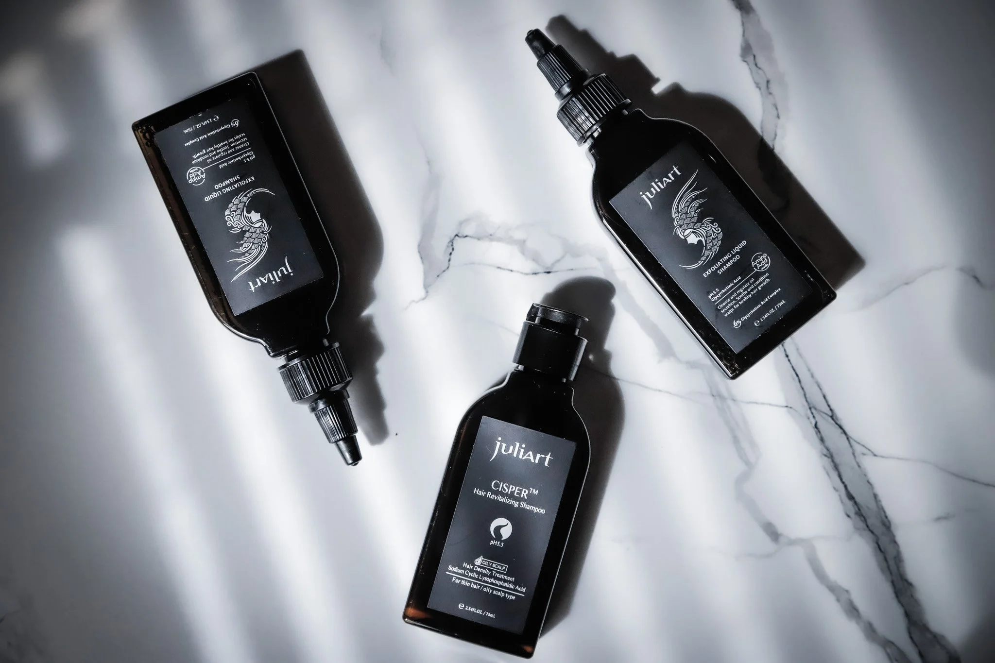 Three black bottles of Juliart hair care products placed on a white marble surface. Two bottles have spray tops, and one has a flip cap. The labels feature a stylized fish illustration and product information.