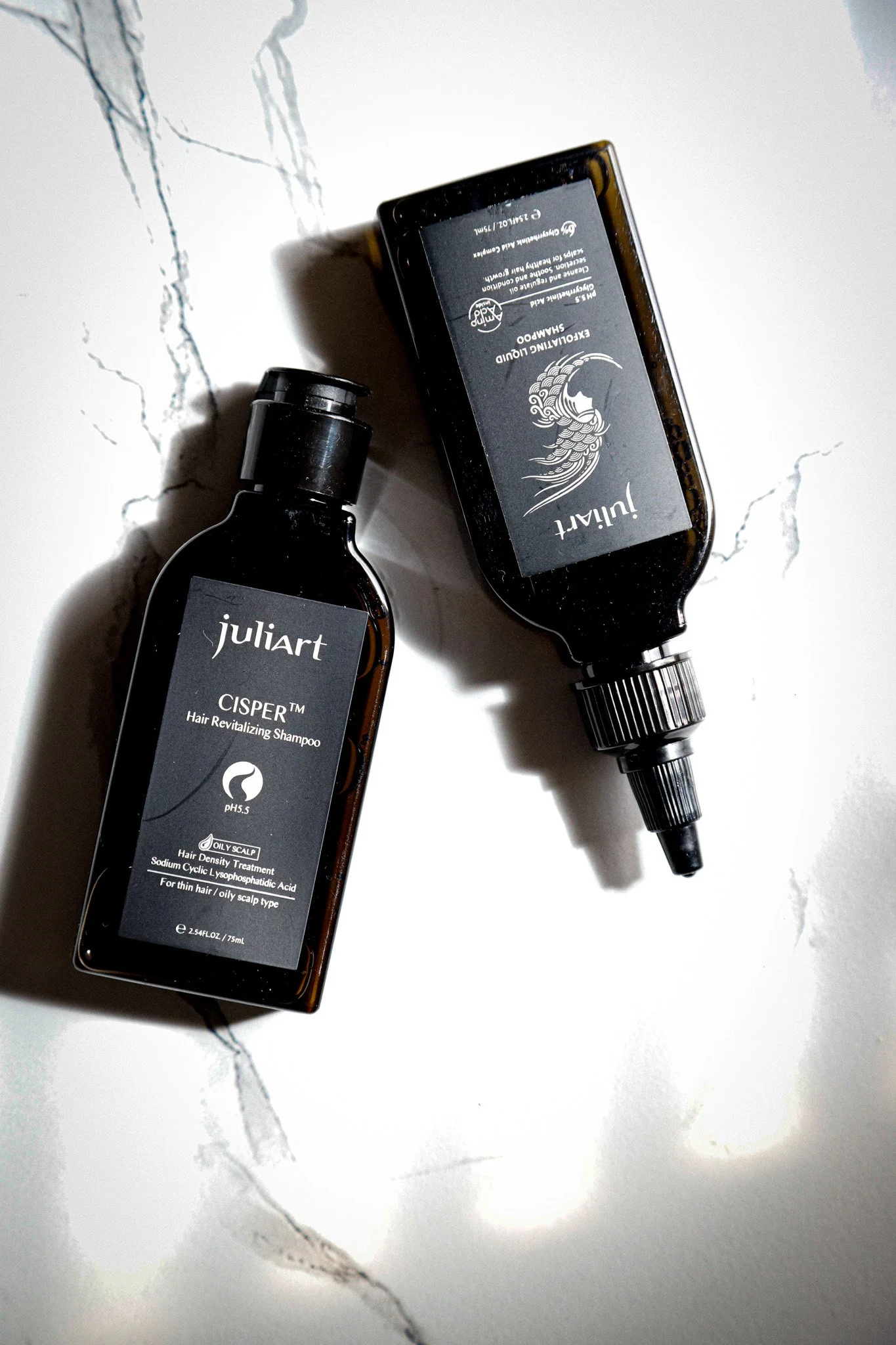 A bottle of Juliart Cisper Hair Revitalizing Shampoo and a bottle of Juliart Exfoliating Liquid are placed on a white marble surface with gray veining.