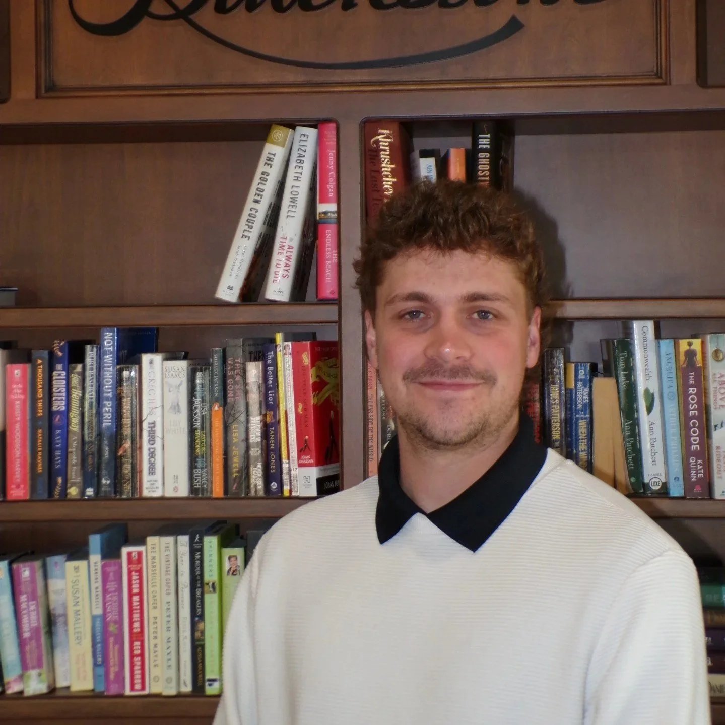 Smiling person in a dress shirt standing inside a room with a stone wall and a large clock in the background.