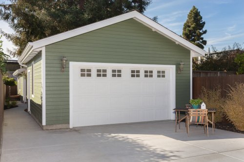 Green single-car garage with white door and windows, outdoor furniture set with a plant, paved driveway, and wooden fence, surrounded by trees.