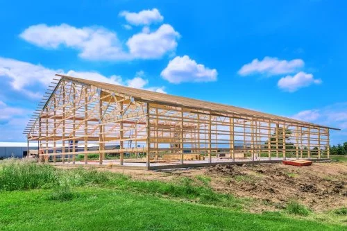 Partially constructed wooden building frame on a grassy field under a blue sky.