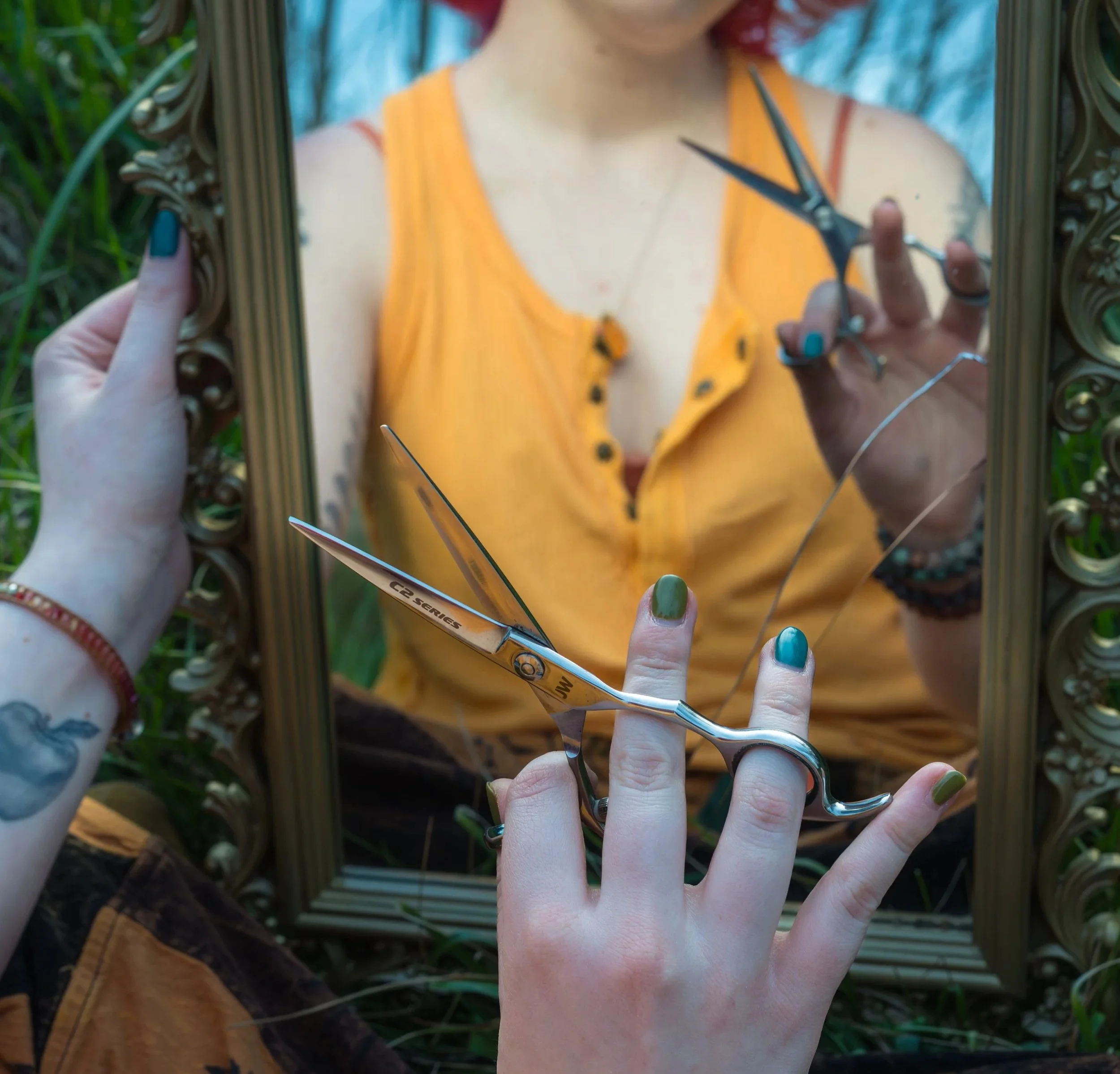 Close-up of a person with colorful nails holding a pair of scissors and cutting a mirror, reflecting their upper body in a yellow top with black buttons and a necklace, in an outdoor setting with greenery.