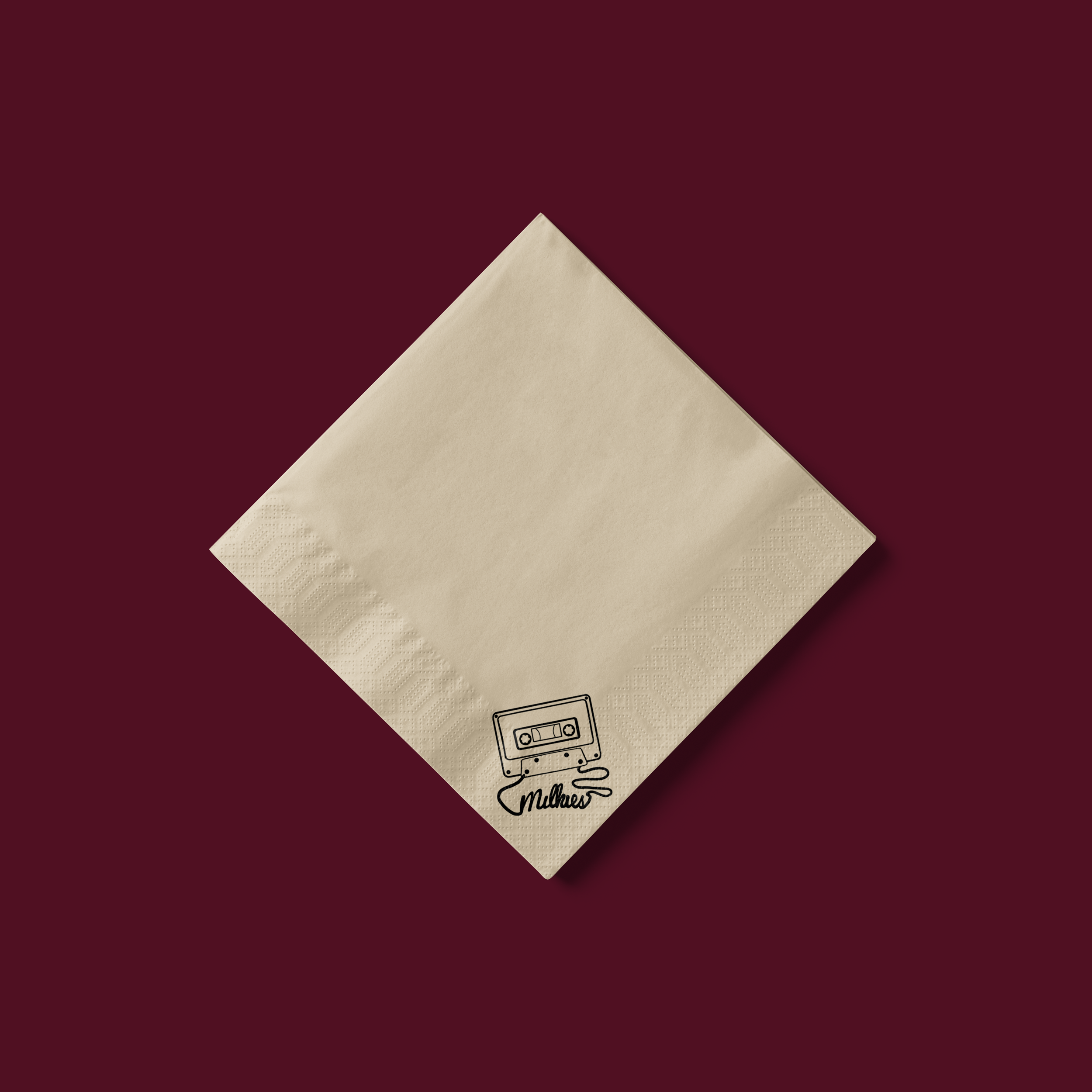 A beige napkin with a black line drawing of a cassette tape and the name 'Milkies' written underneath, placed on a dark maroon surface.