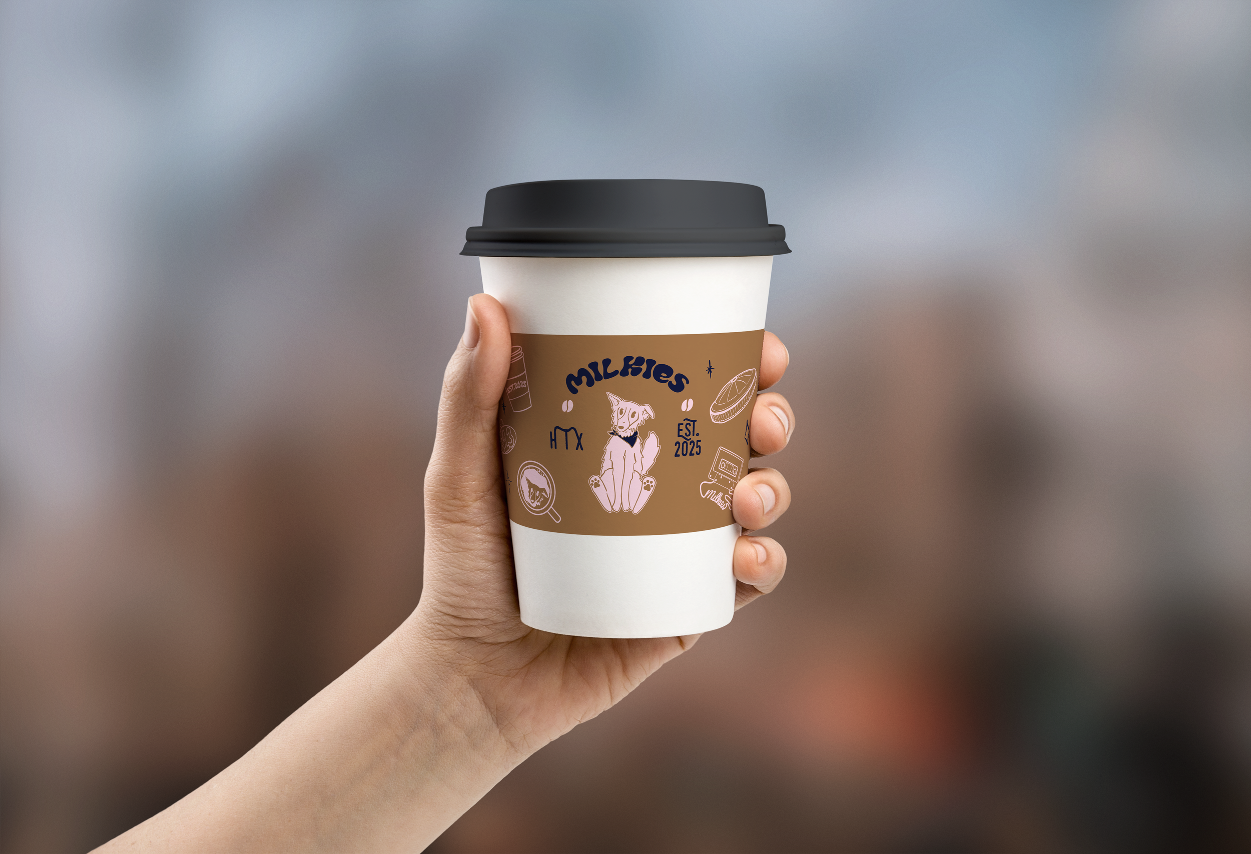 A person holding a disposable coffee cup with a black lid and a paper sleeve featuring the logo of Milkies, a dog illustration, and various food-related icons.