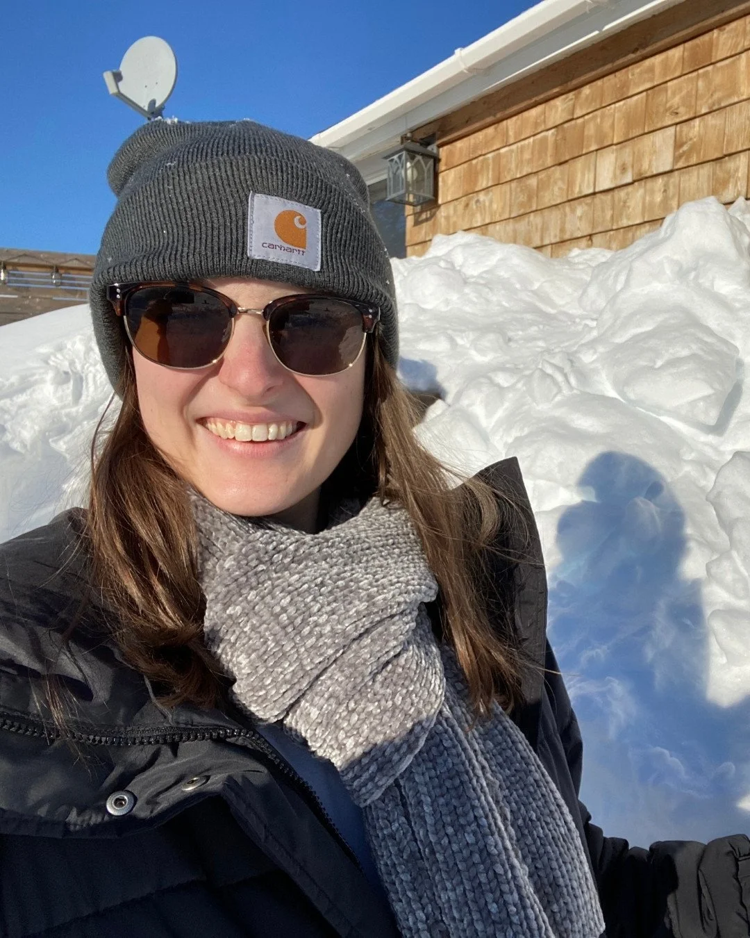A woman smiling outdoors in winter, wearing a gray Carhartt beanie, sunglasses, a gray knitted scarf, and a black winter coat, with snow piled around her and a wooden house behind her.