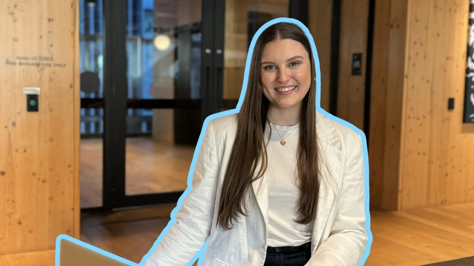 A young woman with long brown hair smiling, wearing a white blazer and necklace, seated indoors with wooden walls and glass doors in the background.