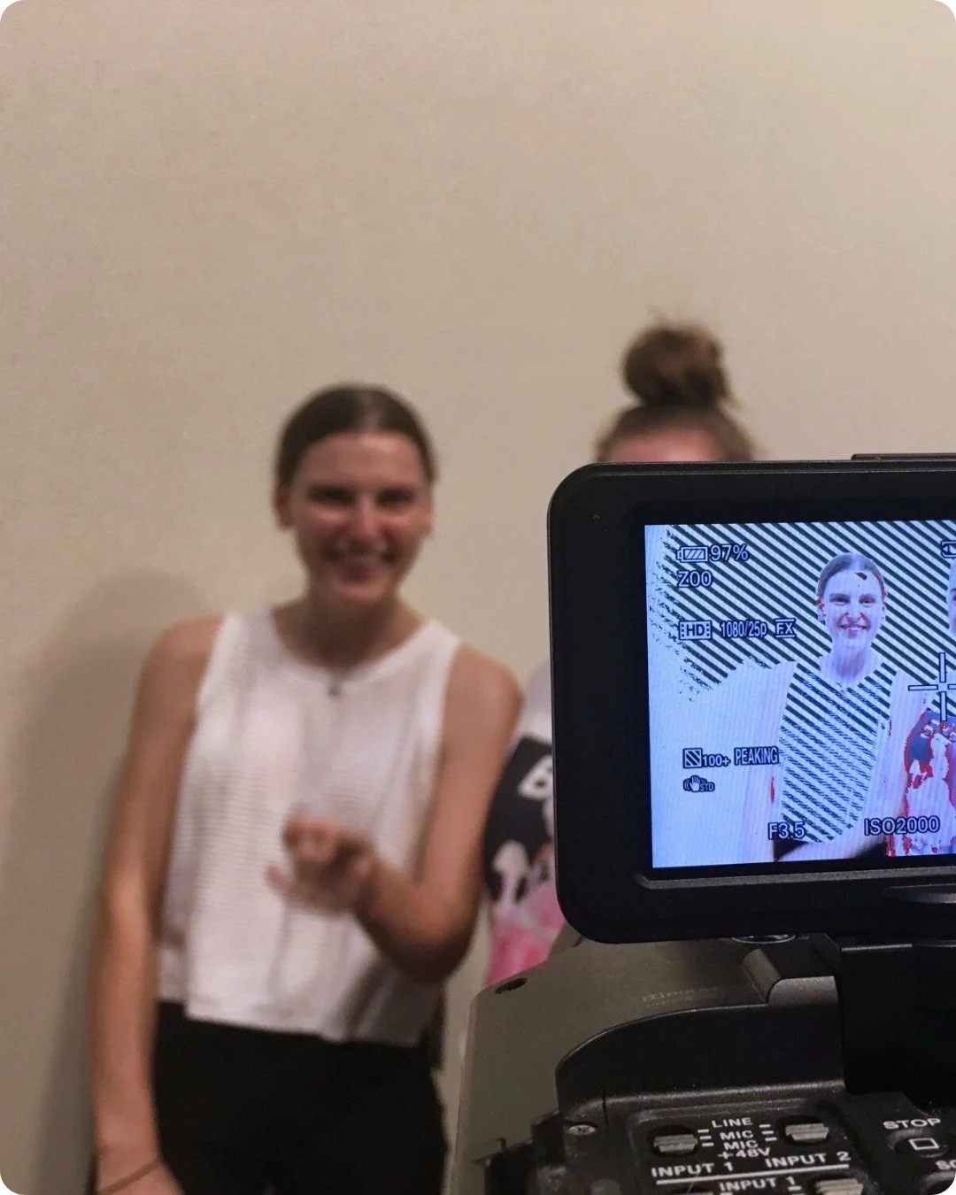 A woman with dark hair smiling and pointing at the camera, with another person partially visible behind her. The photo is being taken with a camera's viewfinder display visible, showing a portrait of her.