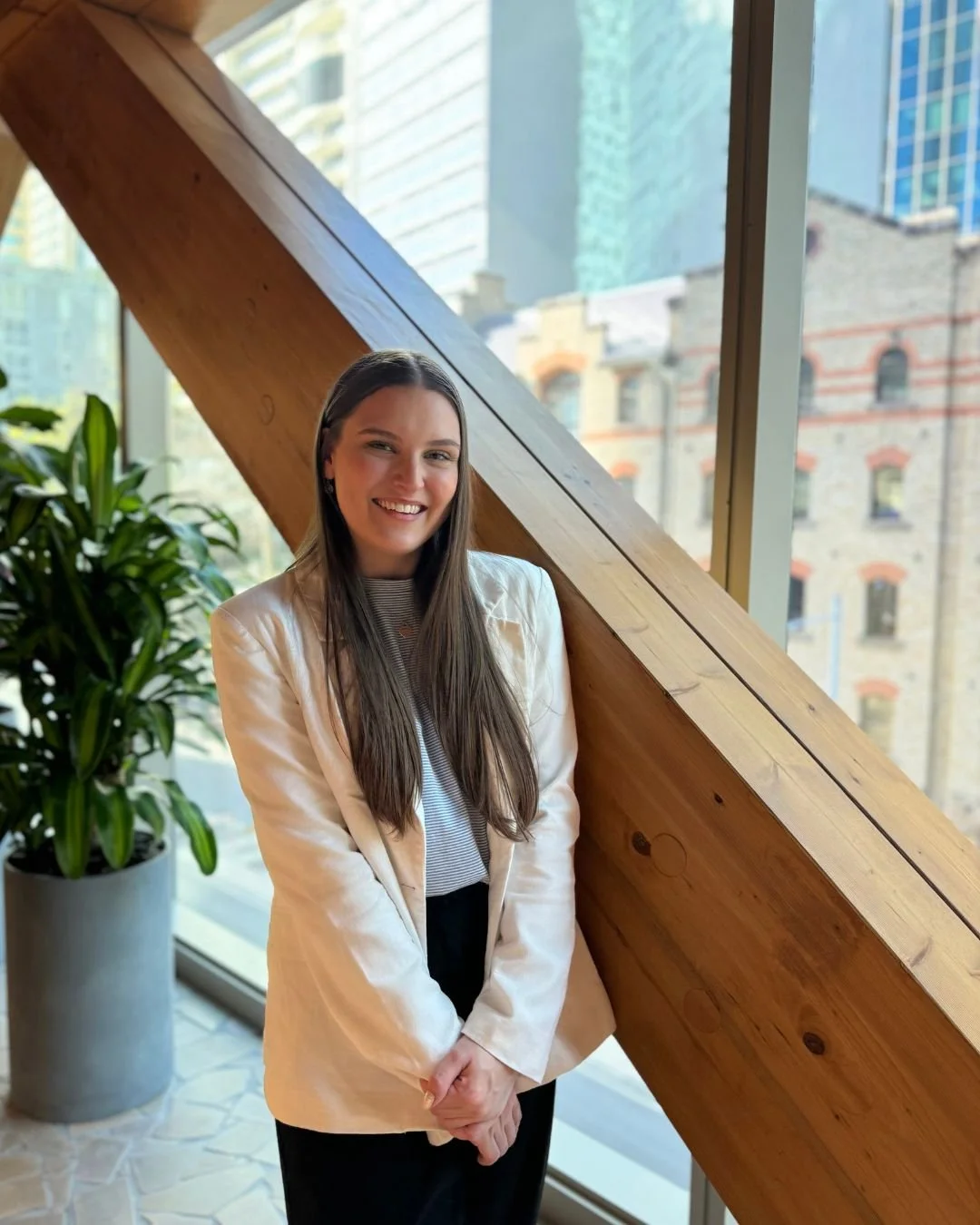 A young woman with long brown hair, wearing a light-colored blazer and black pants, standing indoors near a large window with a view of city buildings, smiling at the camera.