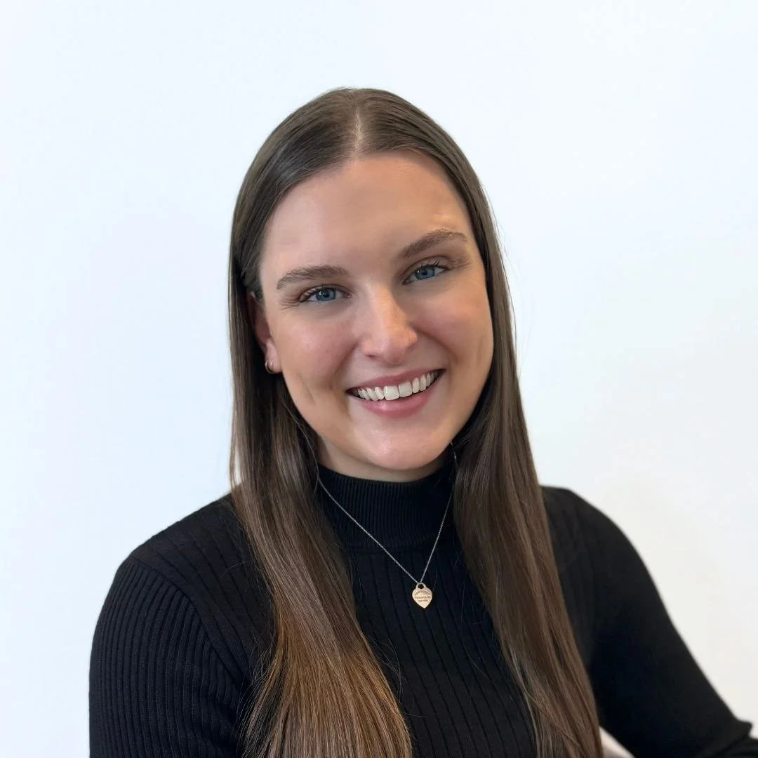 Headshot of a smiling woman with long brown hair in a black turtleneck, wearing a silver necklace and earrings, against a plain white background.