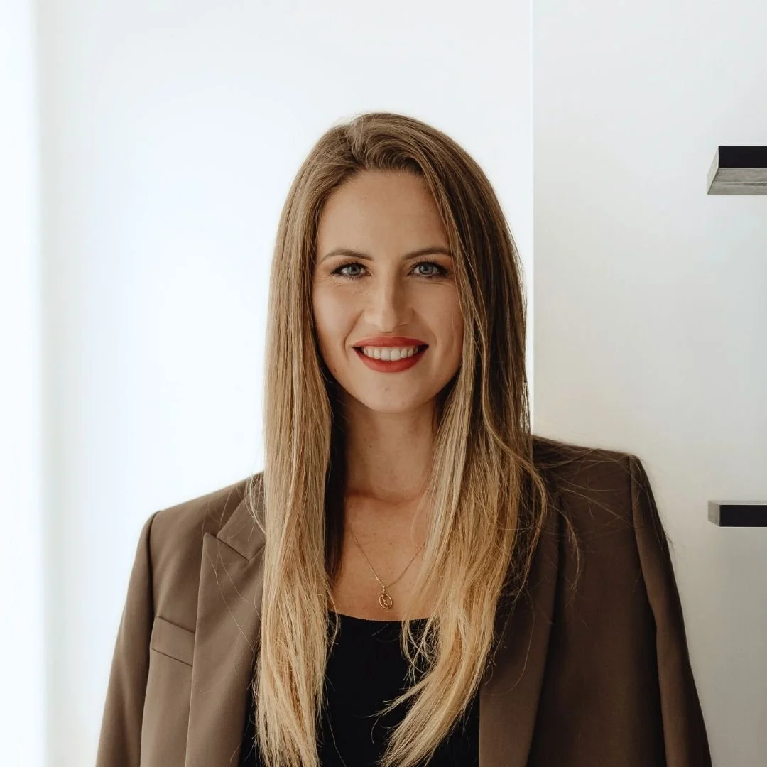 Portrait of a smiling woman with long blonde hair, wearing a dark blazer and a necklace, standing near a white wall.