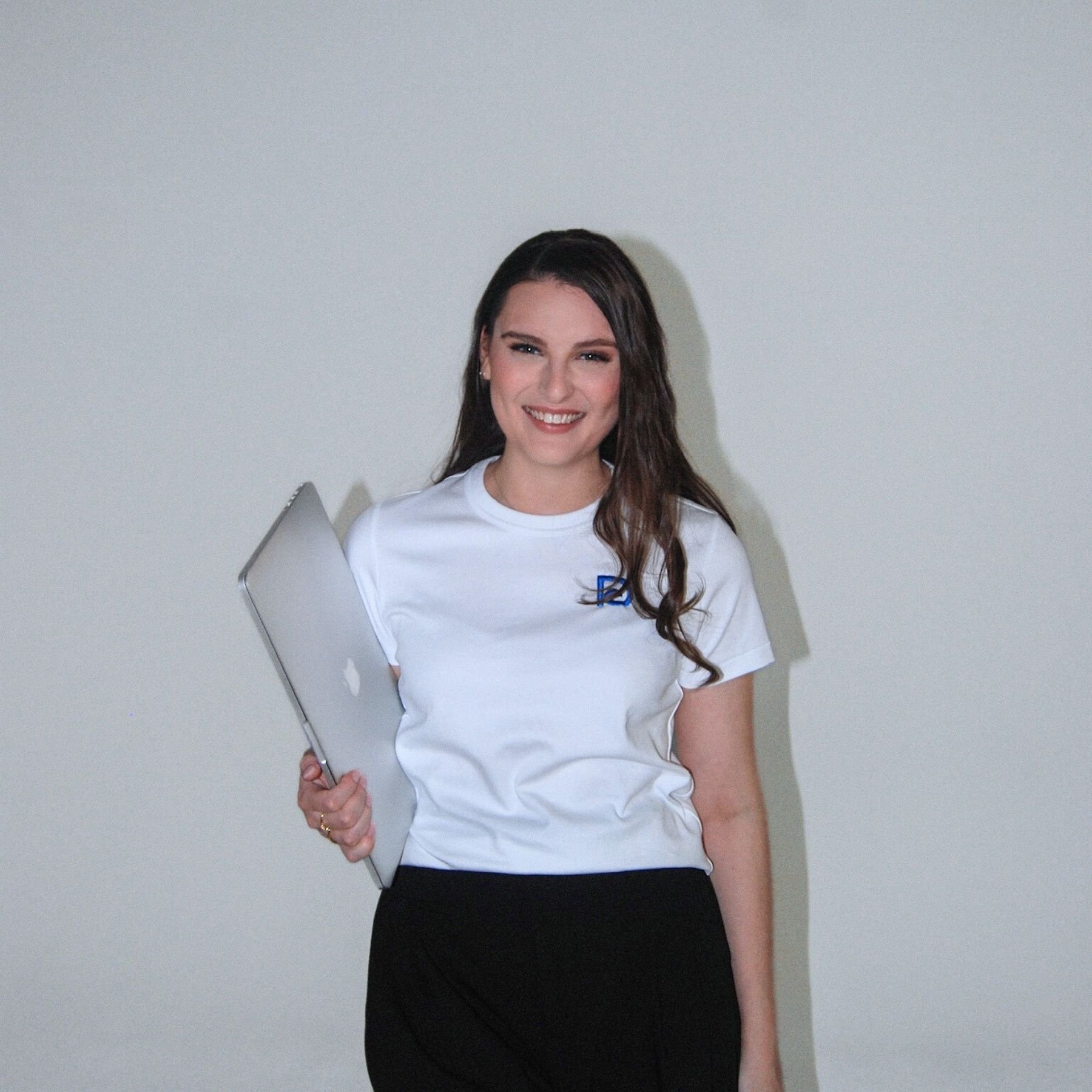 A woman in a white t-shirt holding a silver MacBook laptop, smiling and looking at the camera, standing against a plain white wall.