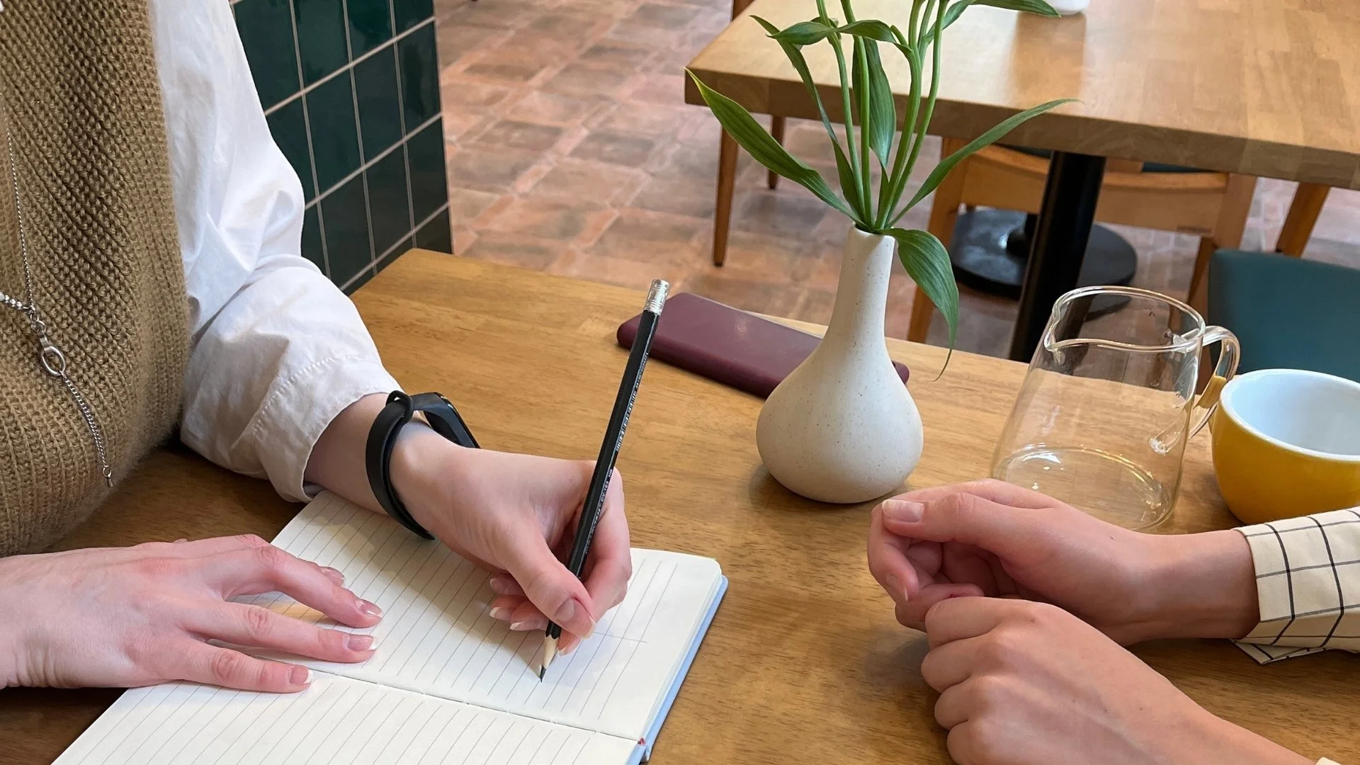 Two people sitting at a wooden table, one taking notes in a lined notebook with a black pencil, and the other resting their hands on the table. On the table are a tall vase with green leaves, a glass pitcher, a yellow mug, and a closed maroon book. The background shows more tables and chairs in a cozy, well-lit space.