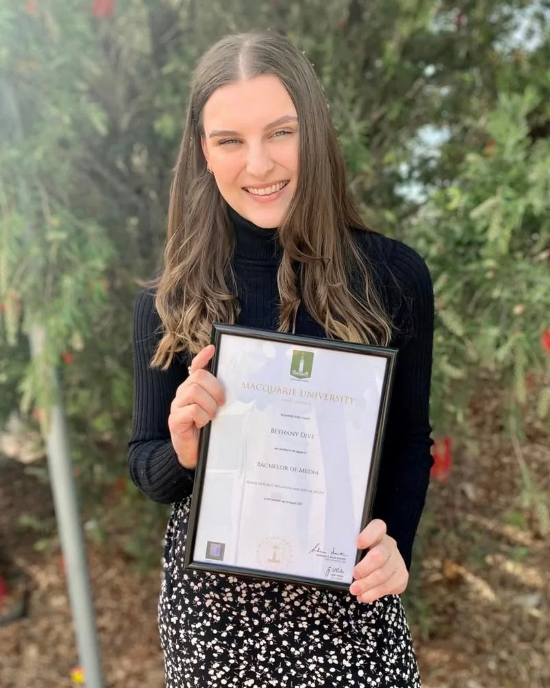 A woman with long brown hair holding a framed university degree. She is smiling and standing outdoors against a background of greenery. The degree is from Macquarie University and she has earned a bachelor's degree in media.
