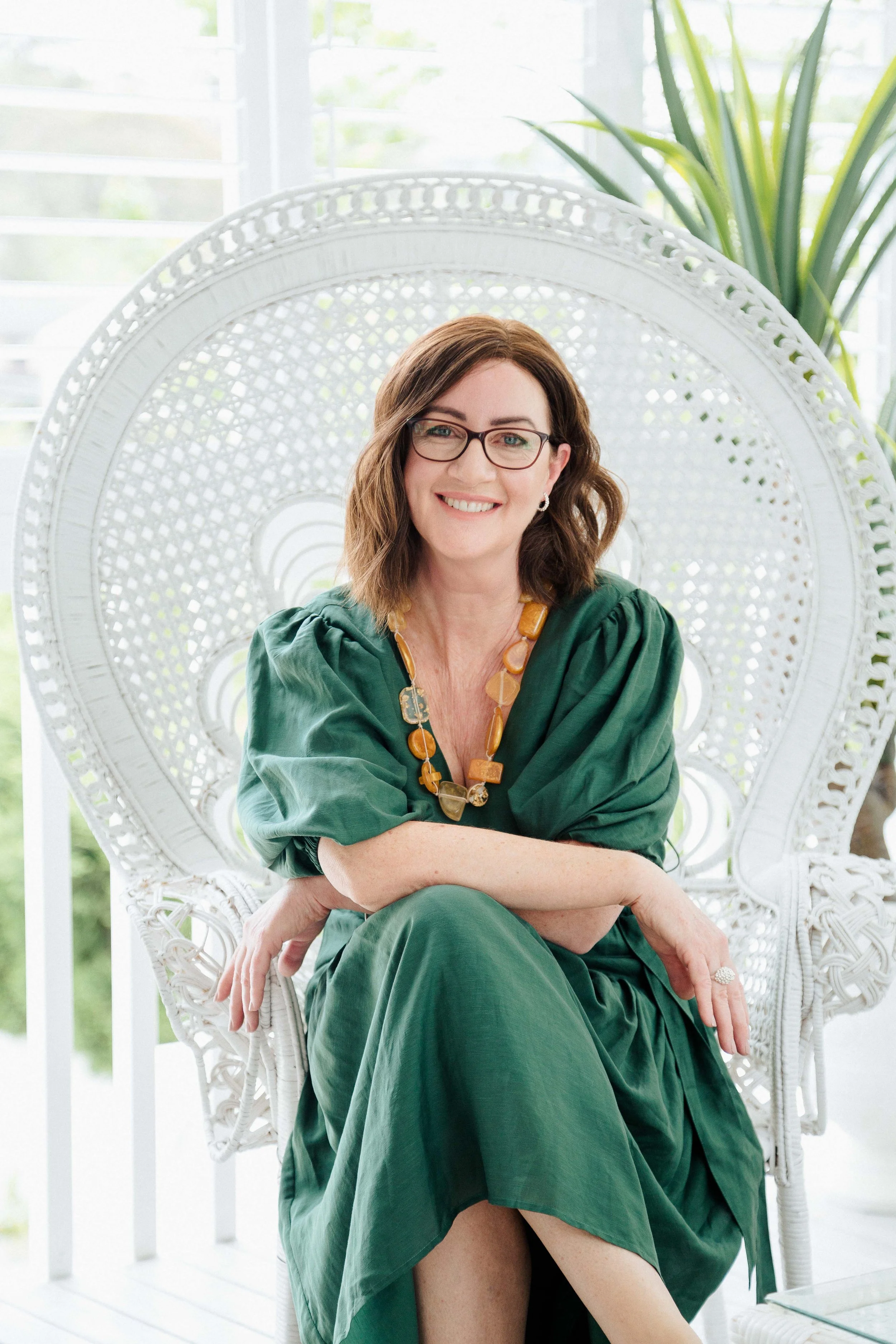 A woman with brown hair, glasses, and a green dress sitting on a white wicker peacock chair, smiling, with green plants and sunlight in the background.