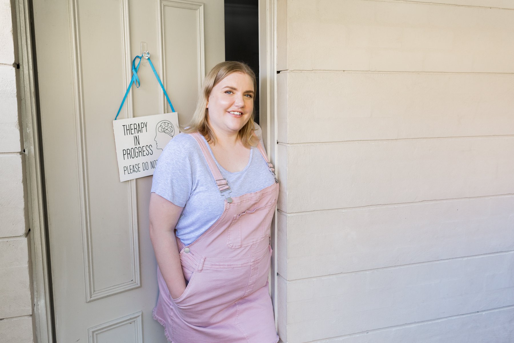 female sex therapist standing in doorway to her clinic wearing a pink dress and grey tee
