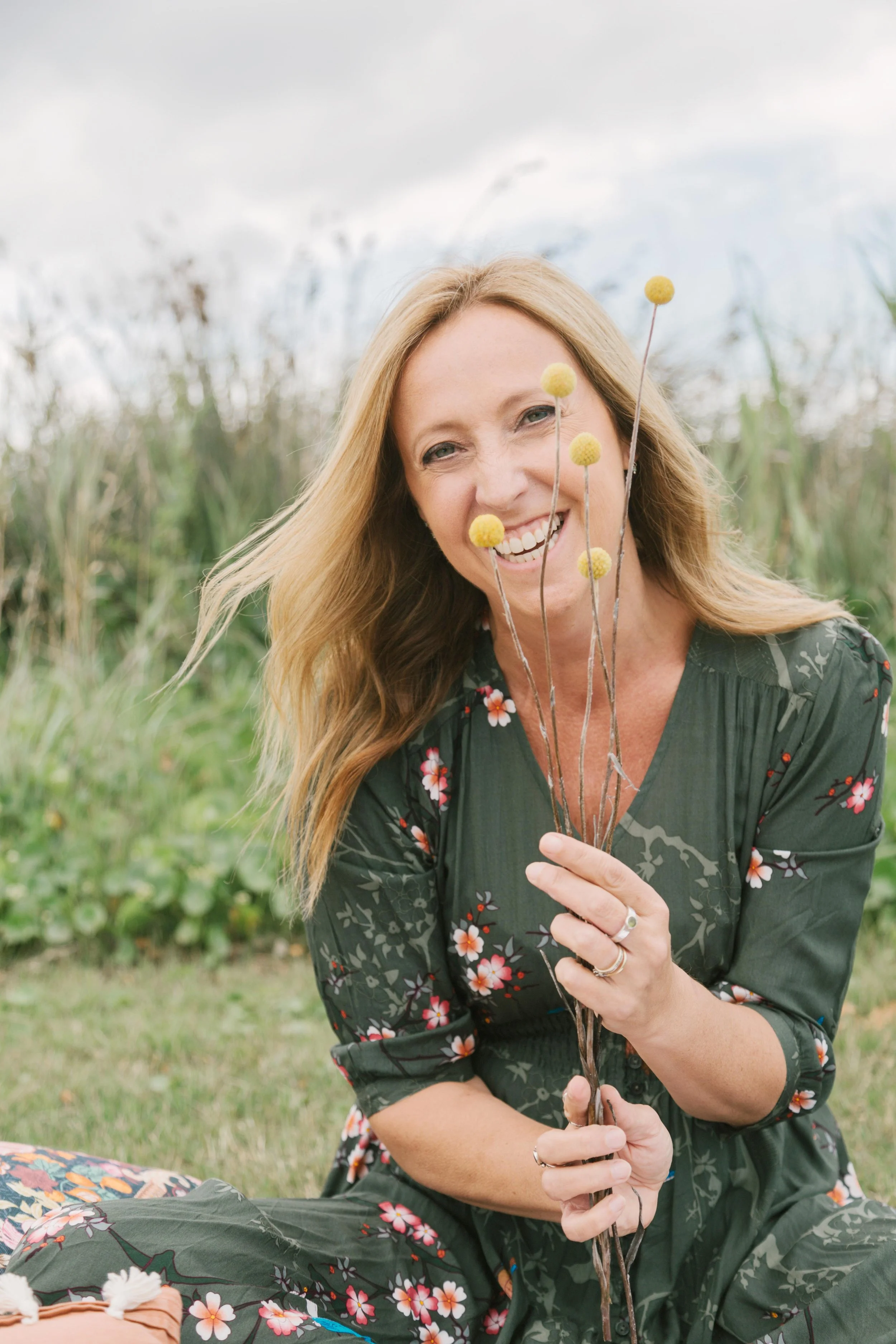blonde woman smiling sitting in a green field holding a small bunch of yellow flowers