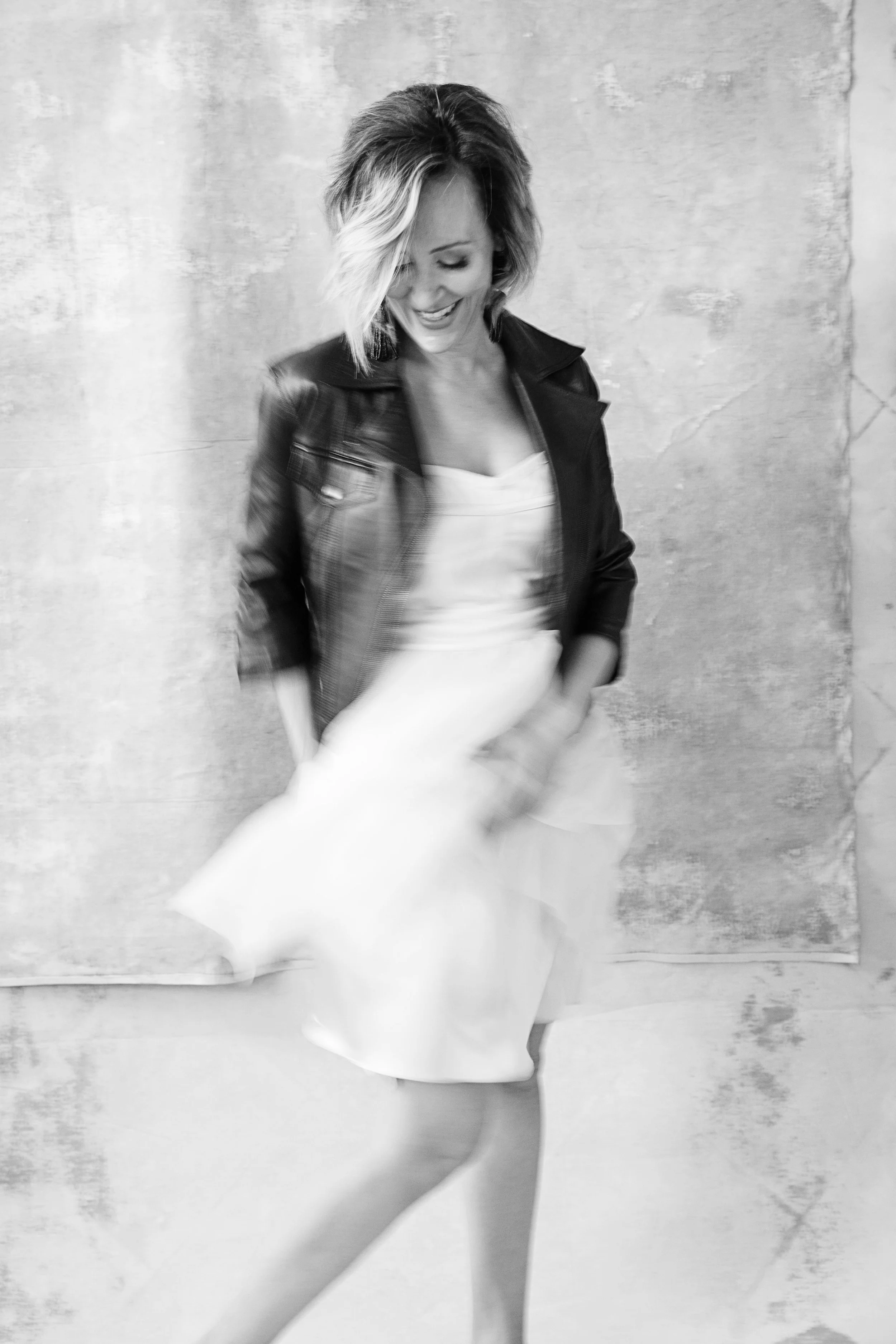 Black and white photo of a woman smiling, wearing a leather jacket over a white dress, standing against a textured wall.