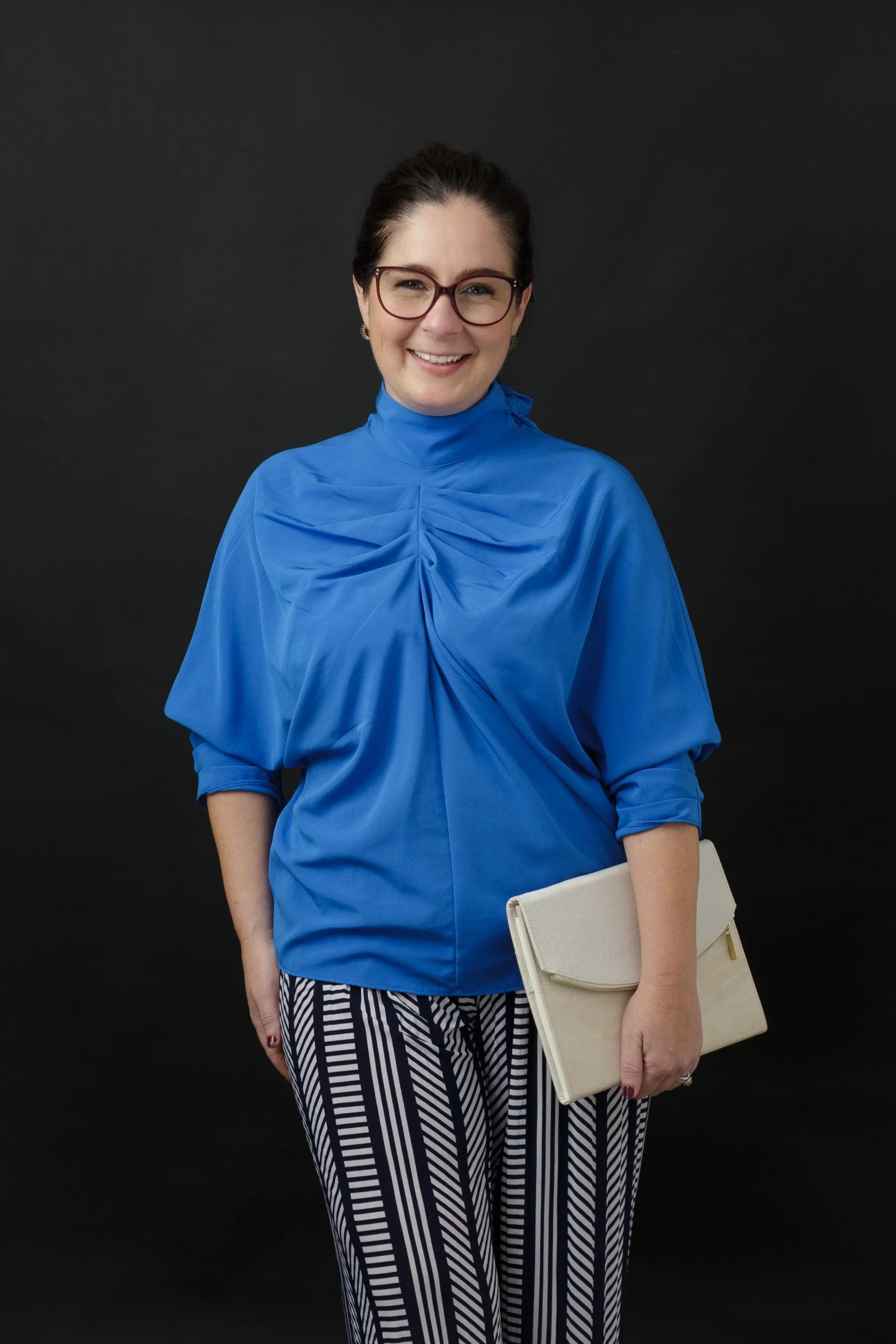 female psychologist with glasses in blue blouse in front of a black backdrop holding a laptop case
