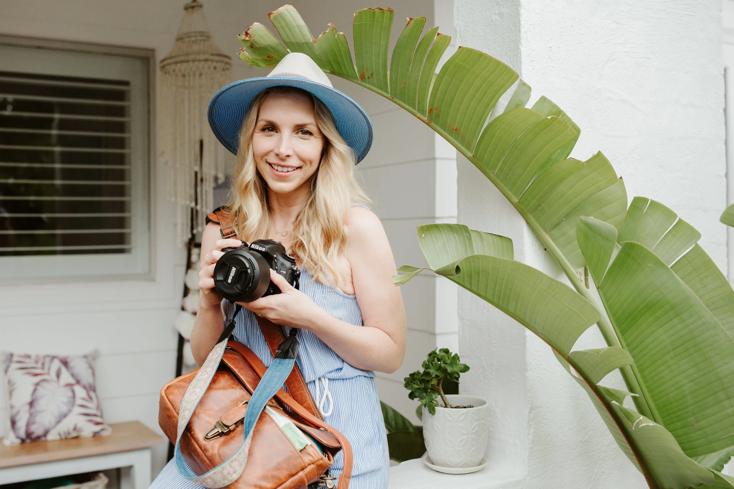 A woman with blonde hair wearing a blue hat and a sleeveless striped dress, holding a camera, standing outside near a large tropical plant, smiling at the camera.