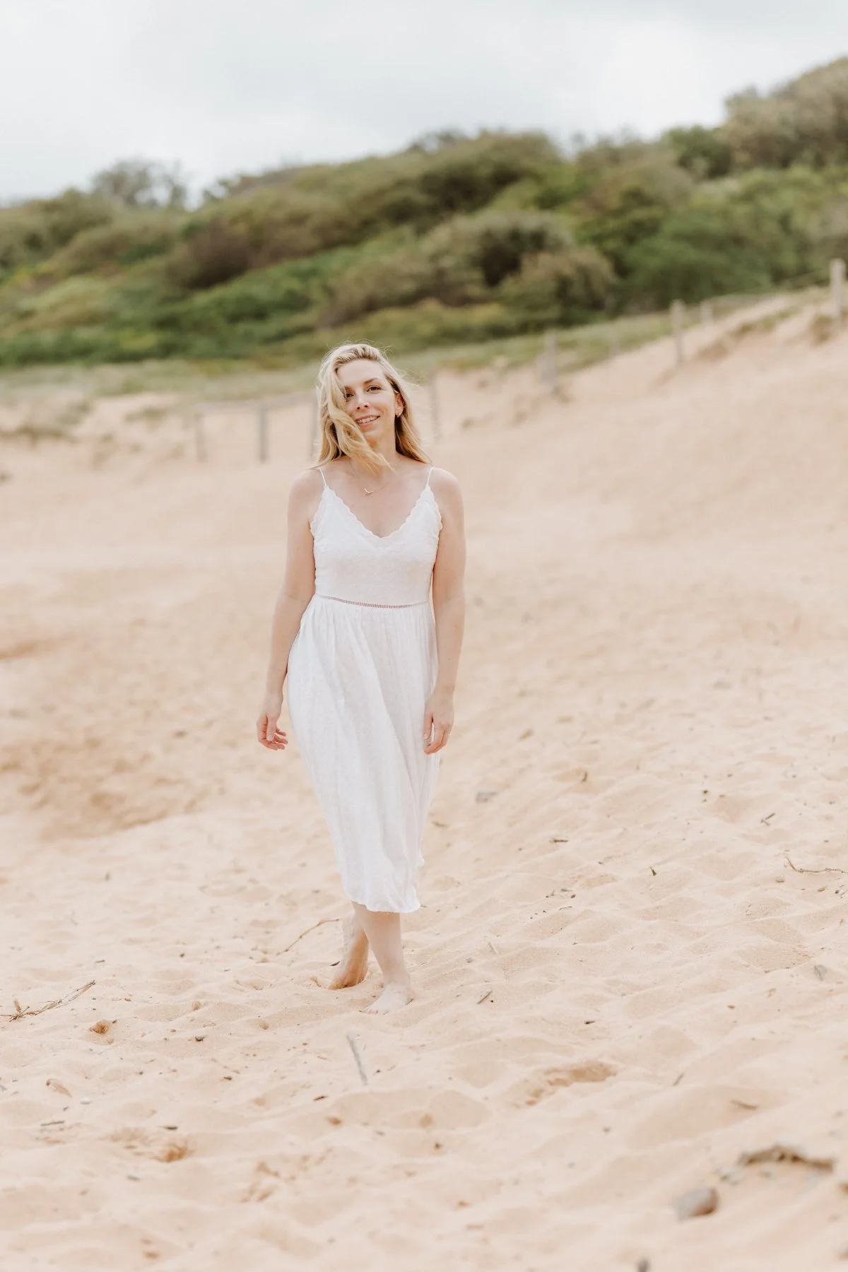 A woman in a white dress walking barefoot on sandy beach with a hill and greenery in the background.
